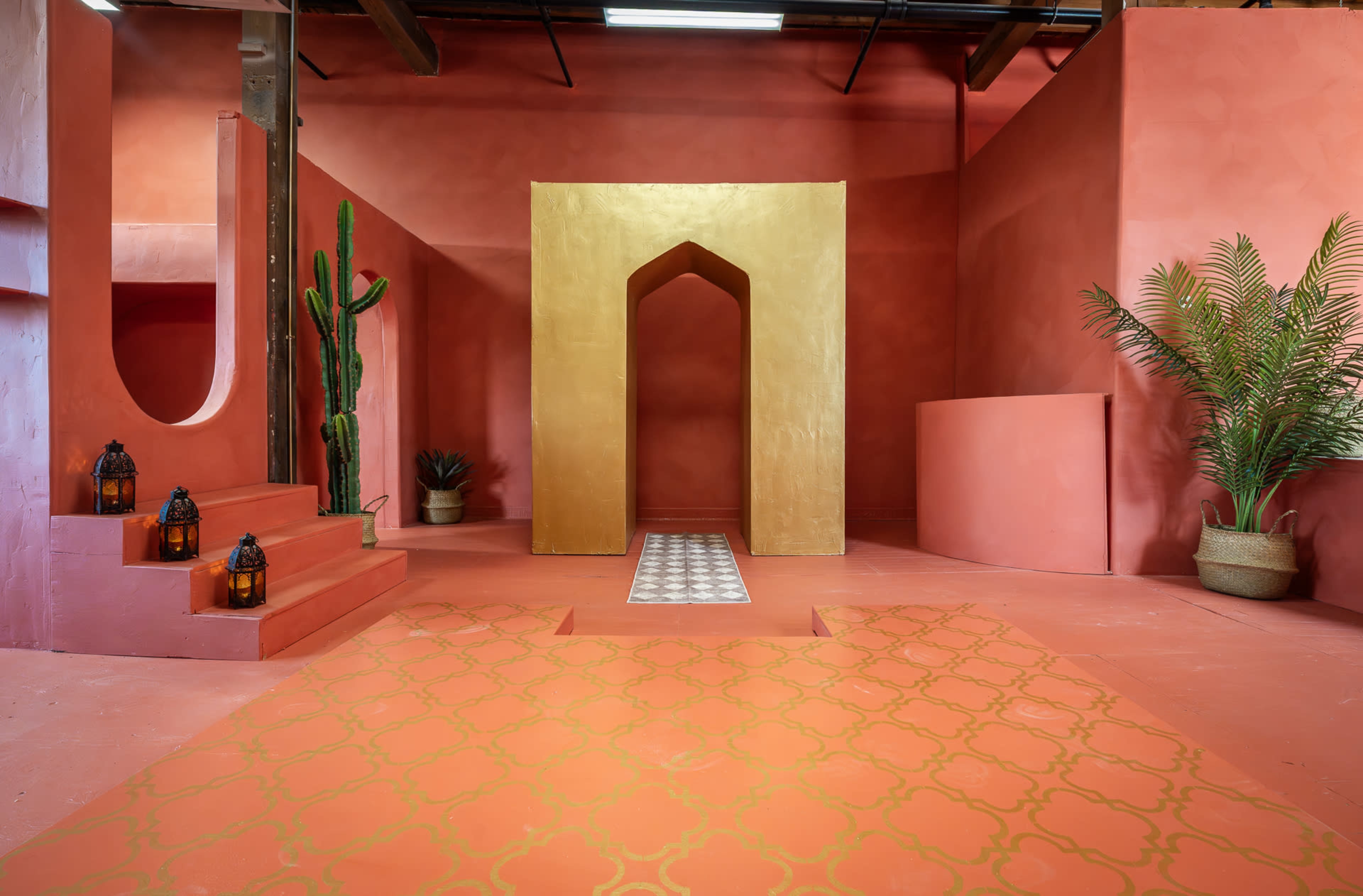 The image depicts a vibrant interior space with pink walls, geometric structures, potted plants, and decorative lanterns arranged around a golden archway and patterned floor.