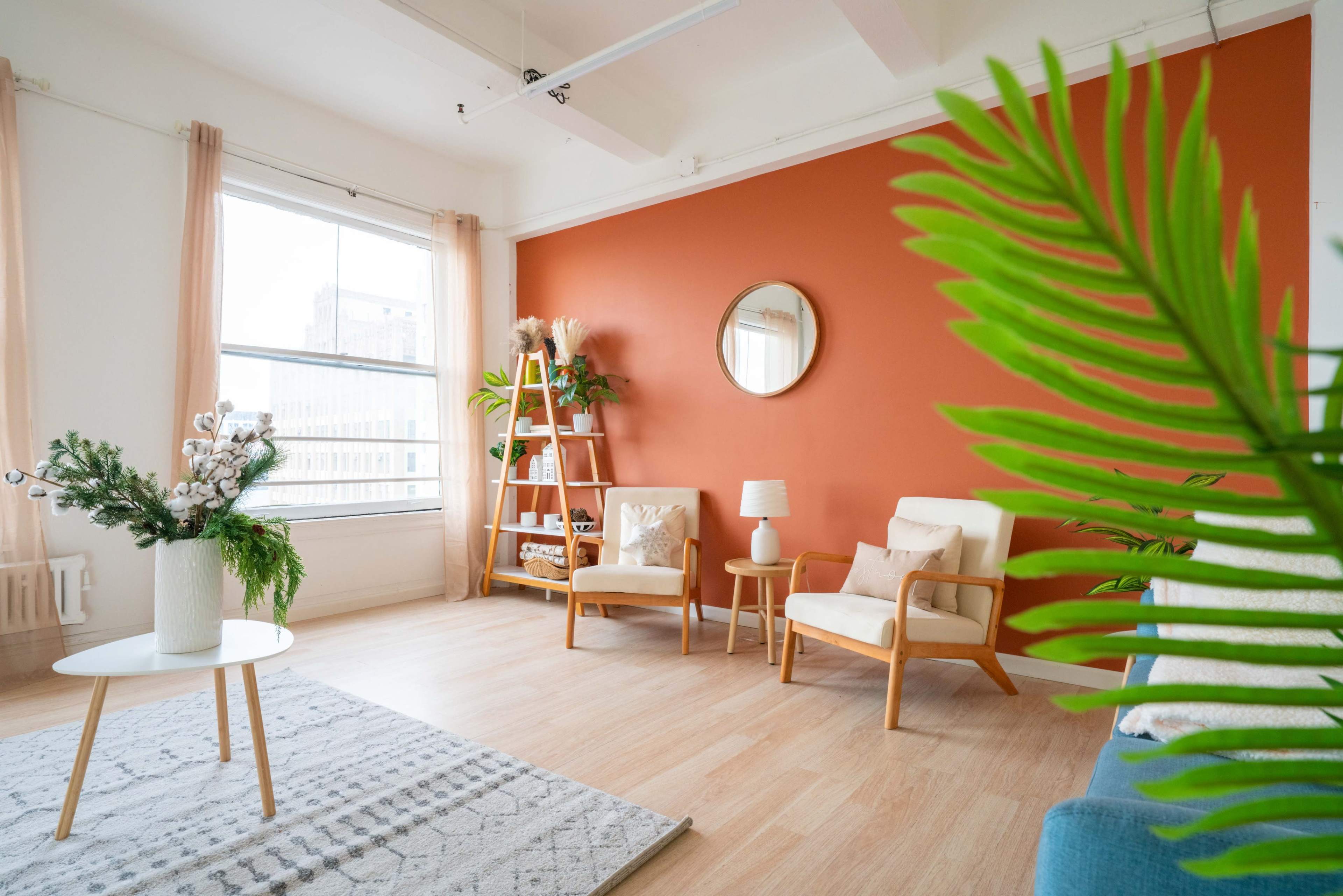 A bright living room featuring a large window, a circular mirror on an orange accent wall, and two light-colored chairs next to a small table, with decorative plants and a shelf in the background.