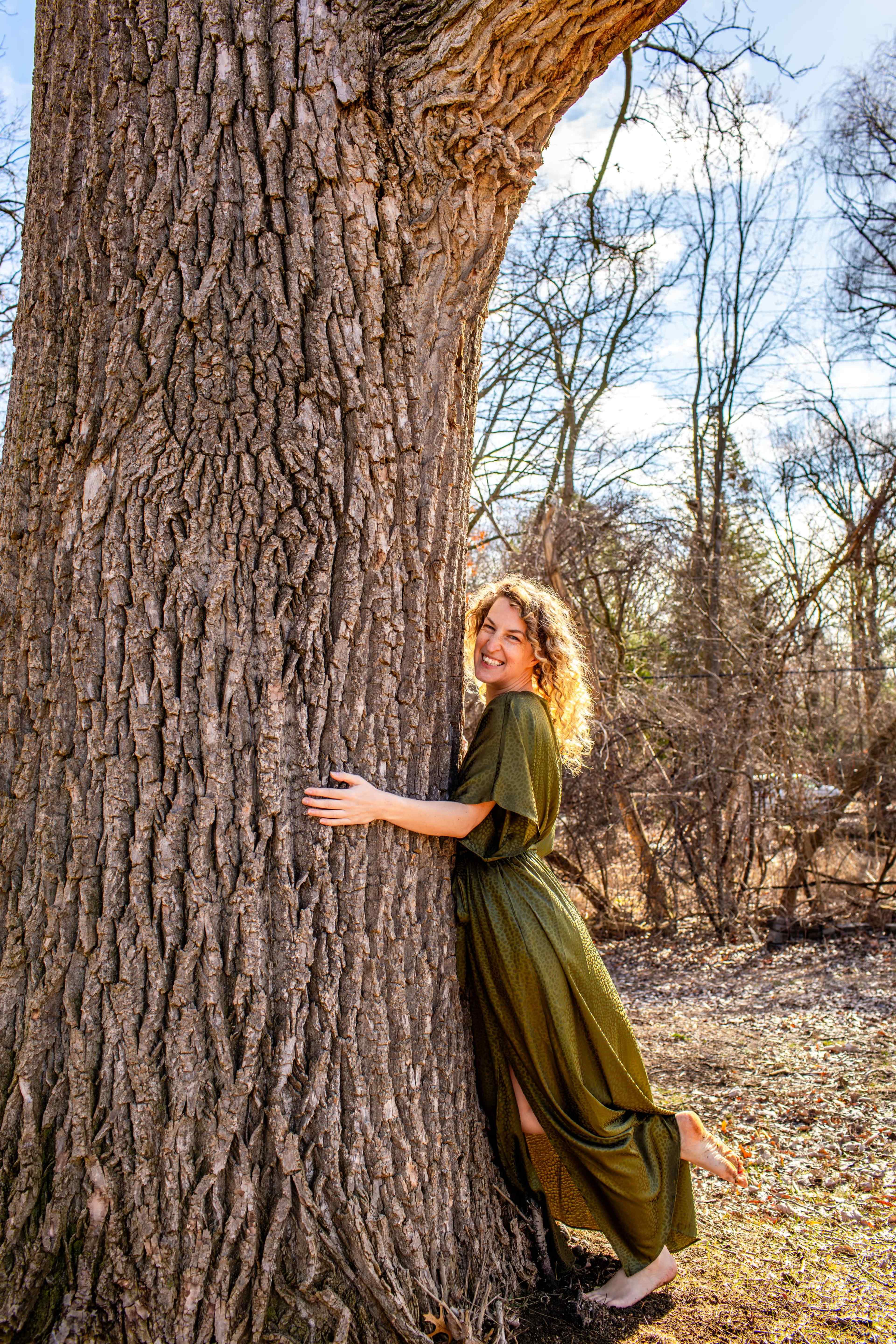 A woman in a long green dress leans against a large tree trunk in a sunlit outdoor setting.