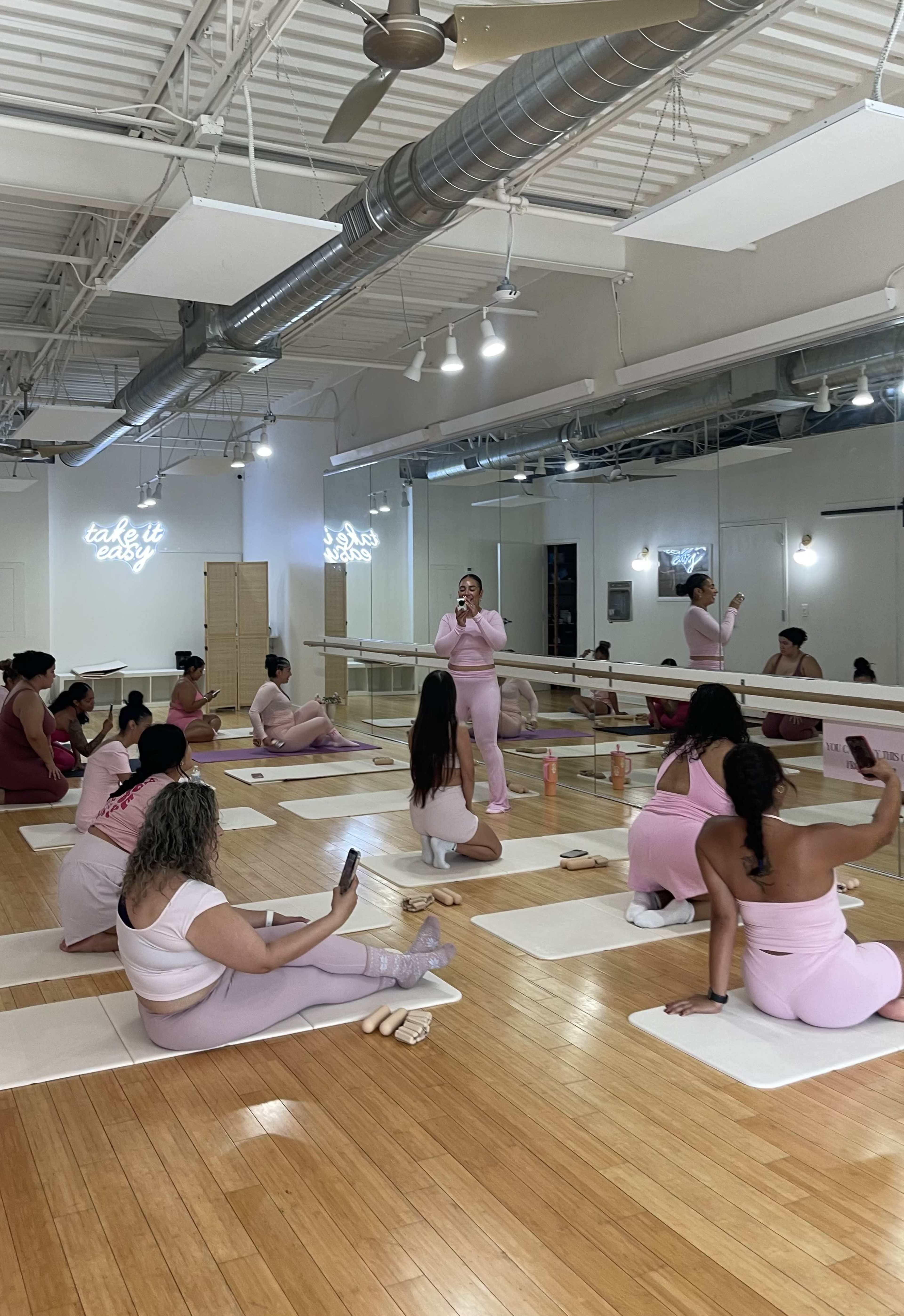 A fitness class is taking place in a studio with participants wearing pink attire, sitting on yoga mats while a coach addresses them.