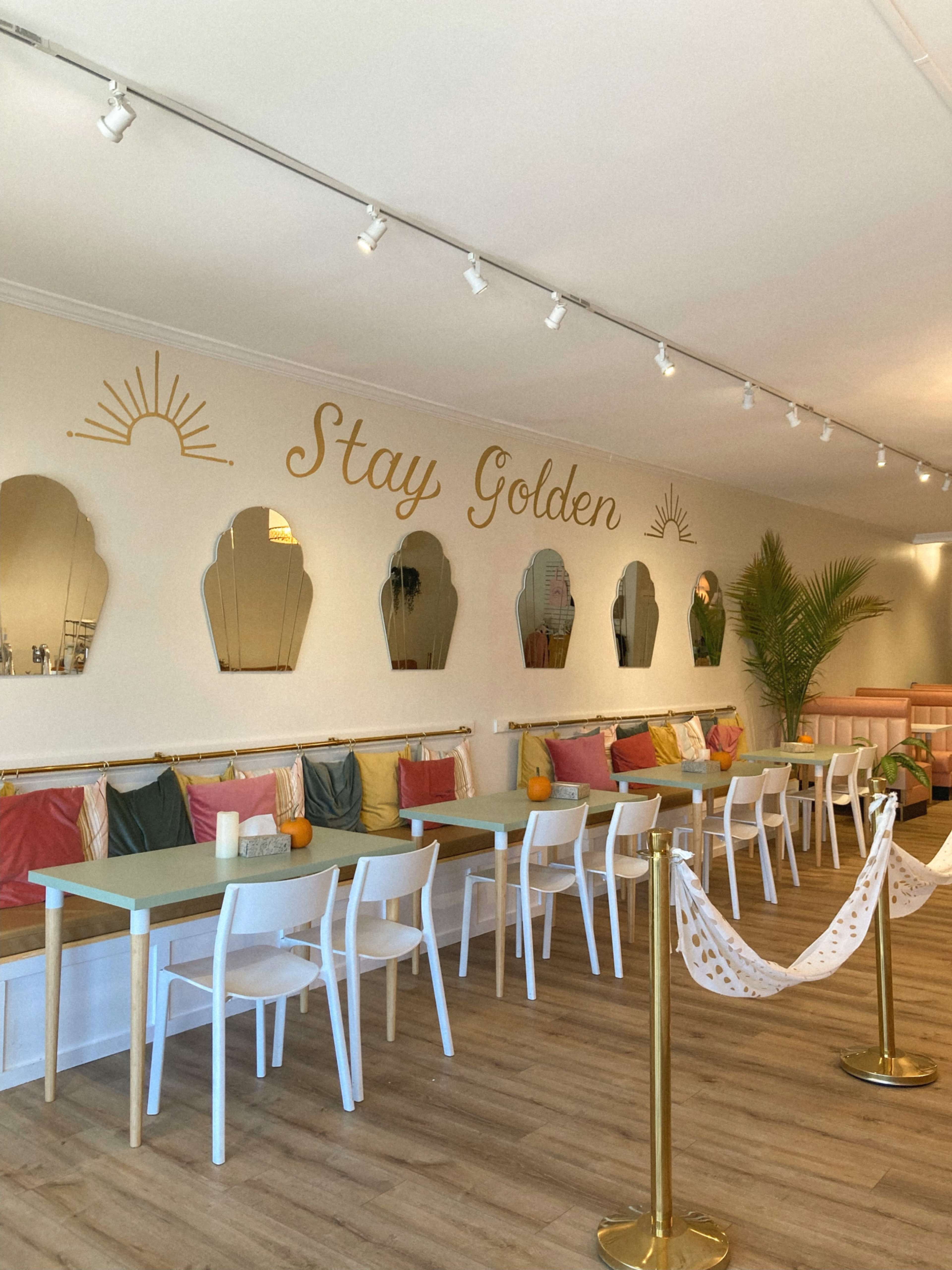A brightly lit café features a white wall with the phrase "Stay Golden" and a row of mirrors above a colorful seating area adorned with cushions.