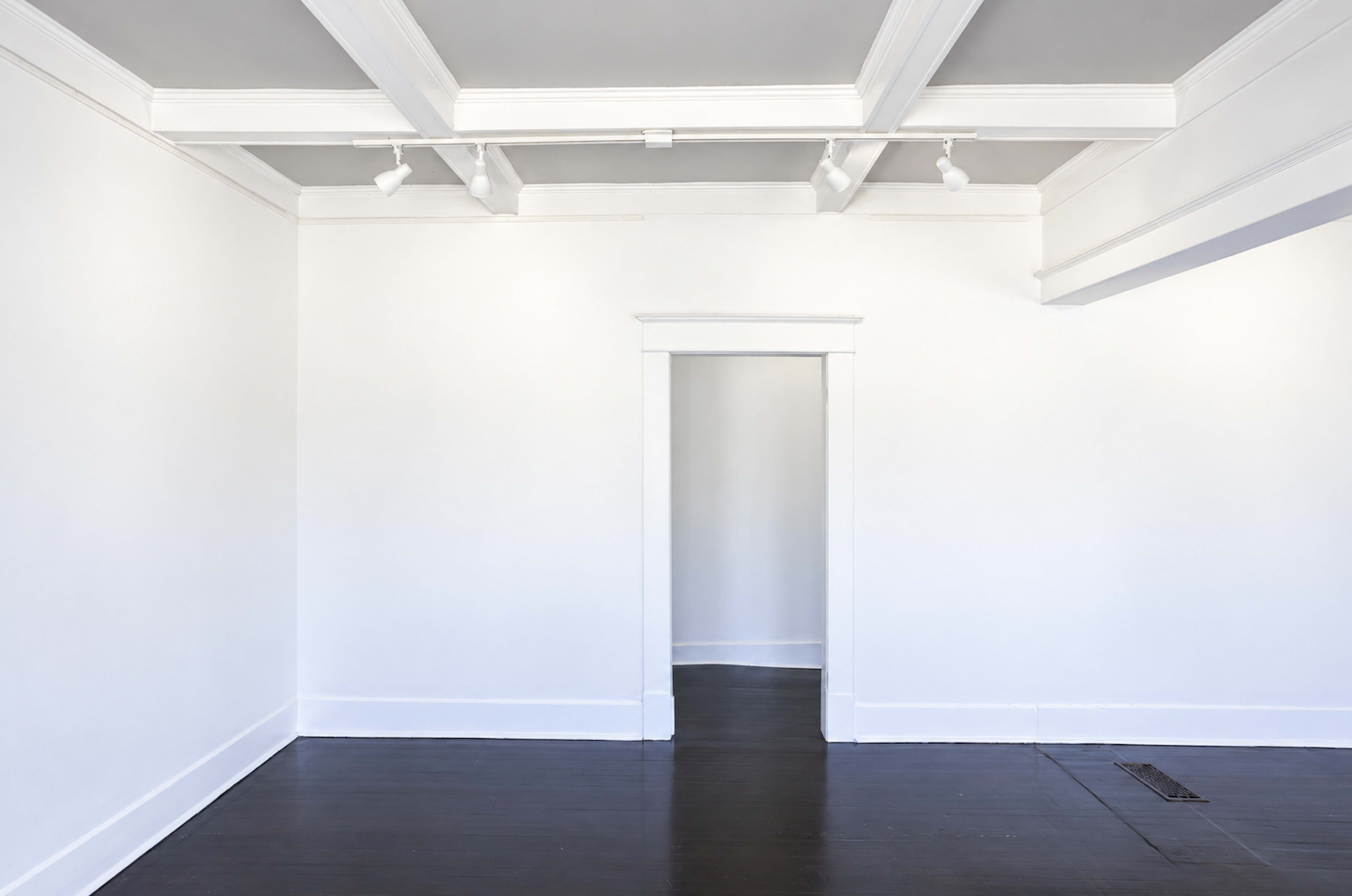 The image shows a sparsely furnished room with white walls, a dark wooden floor, and an open doorway leading to another space.