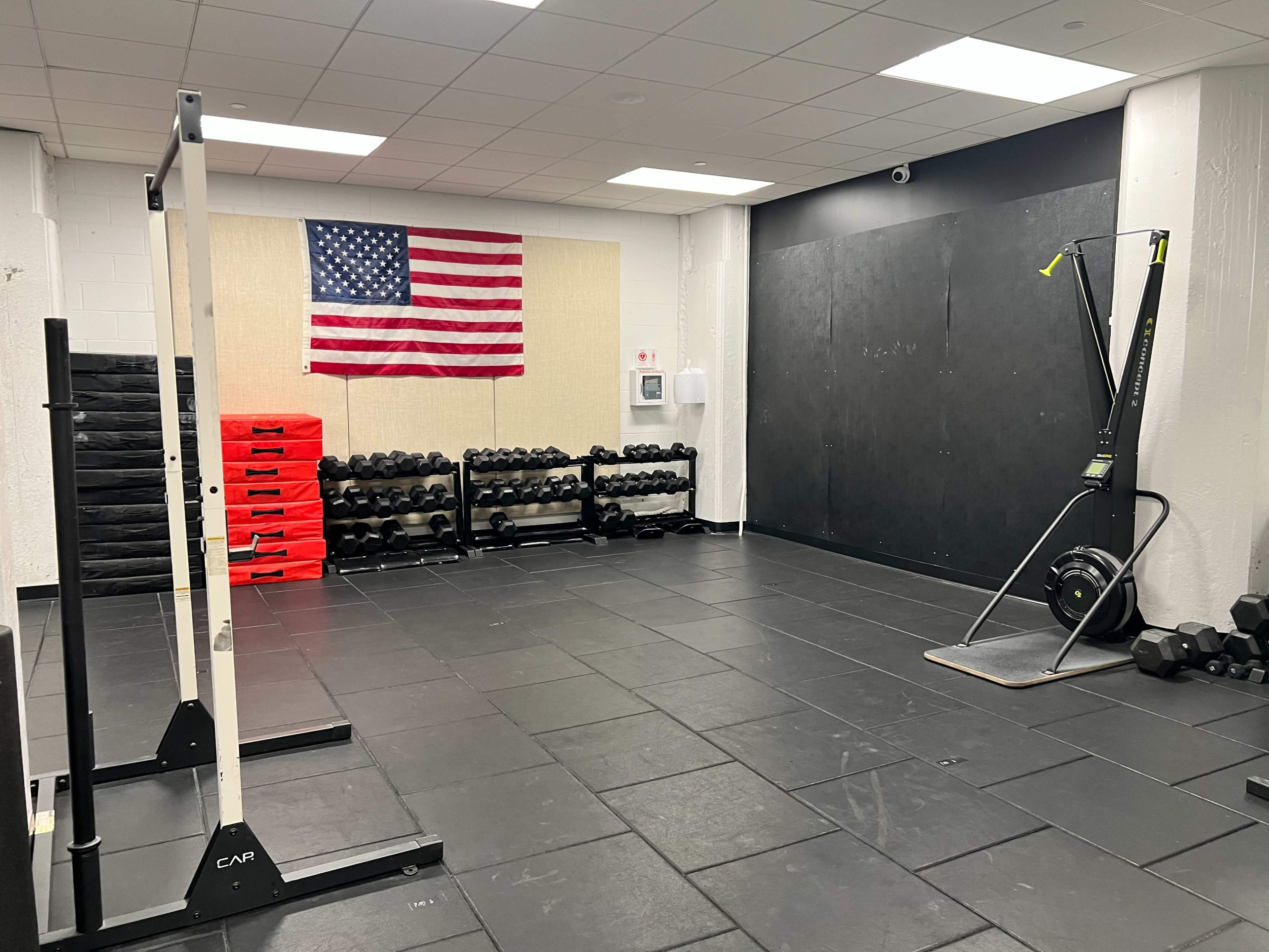 A gym room featuring various fitness equipment, including dumbbells on racks, a rowing machine, and an American flag hanging on the wall.