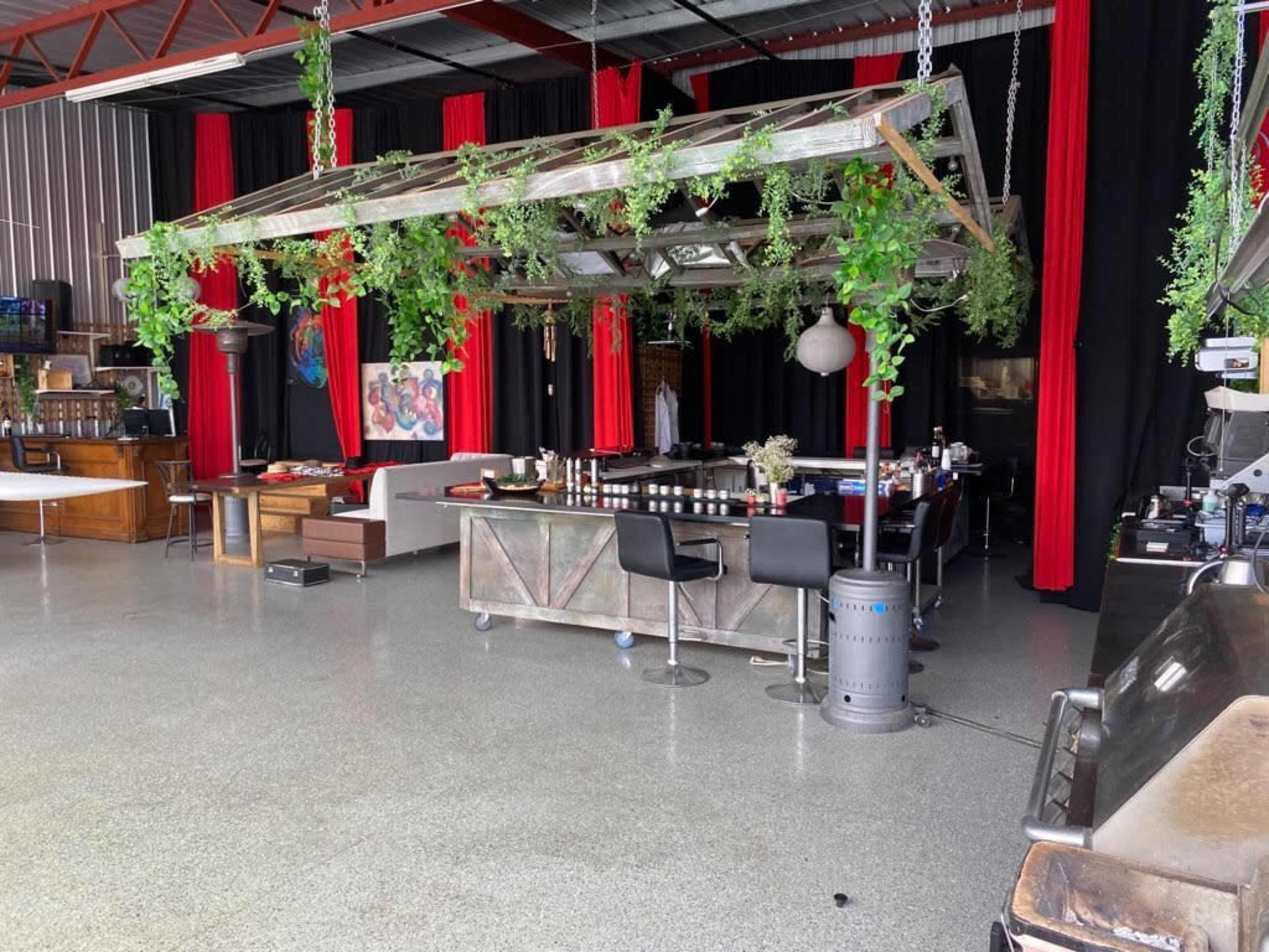 A spacious indoor venue features a bar surrounded by greenery and red curtains, with various seating areas and artistic decor.