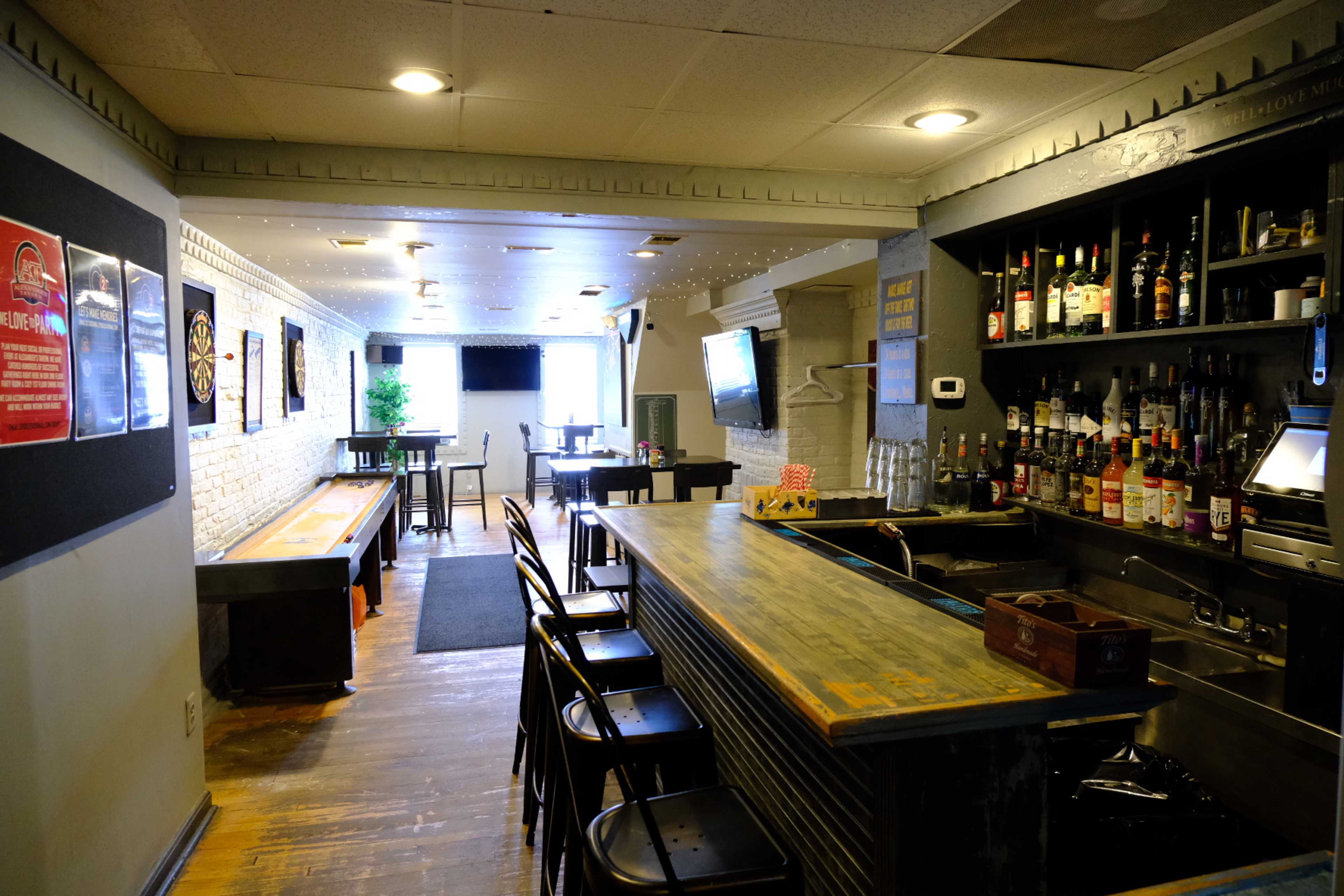 The interior of a bar with a long wooden bar counter, shelves lined with various bottles, a dartboard area, and additional seating in the background.