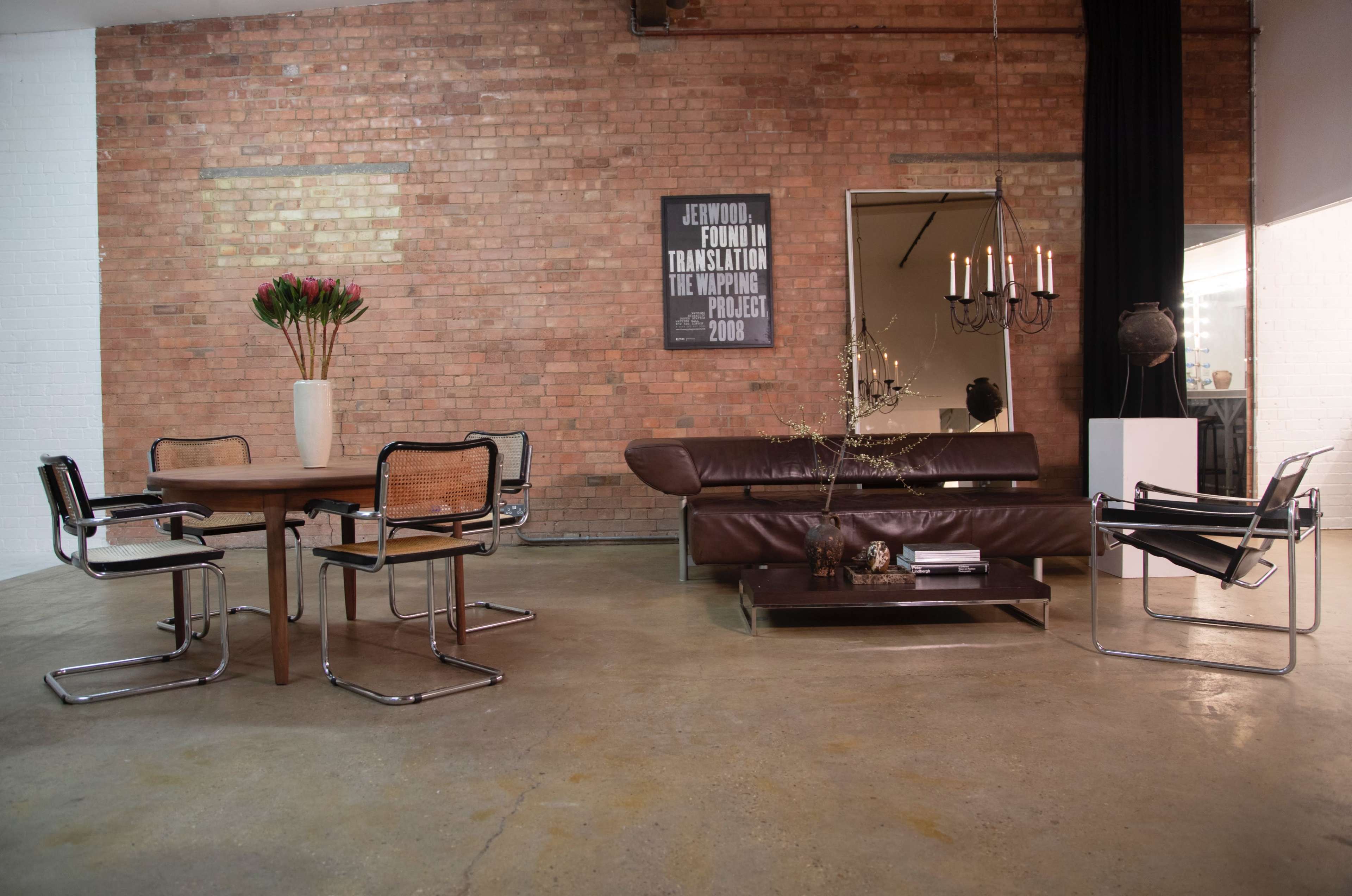 The scene features a minimalist living space with a round wooden dining table, four black and chrome chairs, a brown leather sofa, a large vase with flowers, and a wall-mounted artwork in a room with exposed brick walls.