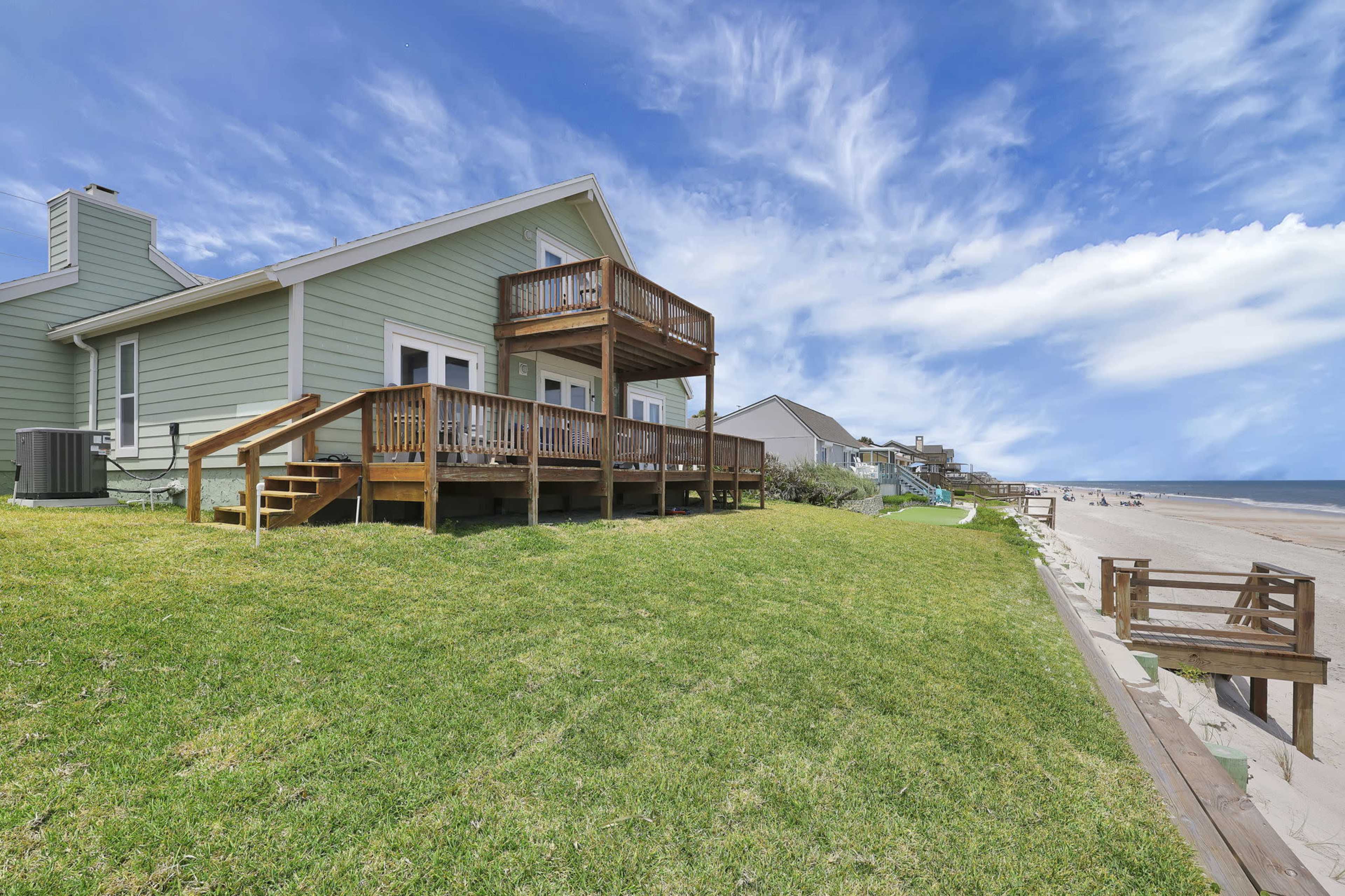 A beach house with a wooden deck overlooks the sandy shore under a partly cloudy sky.
