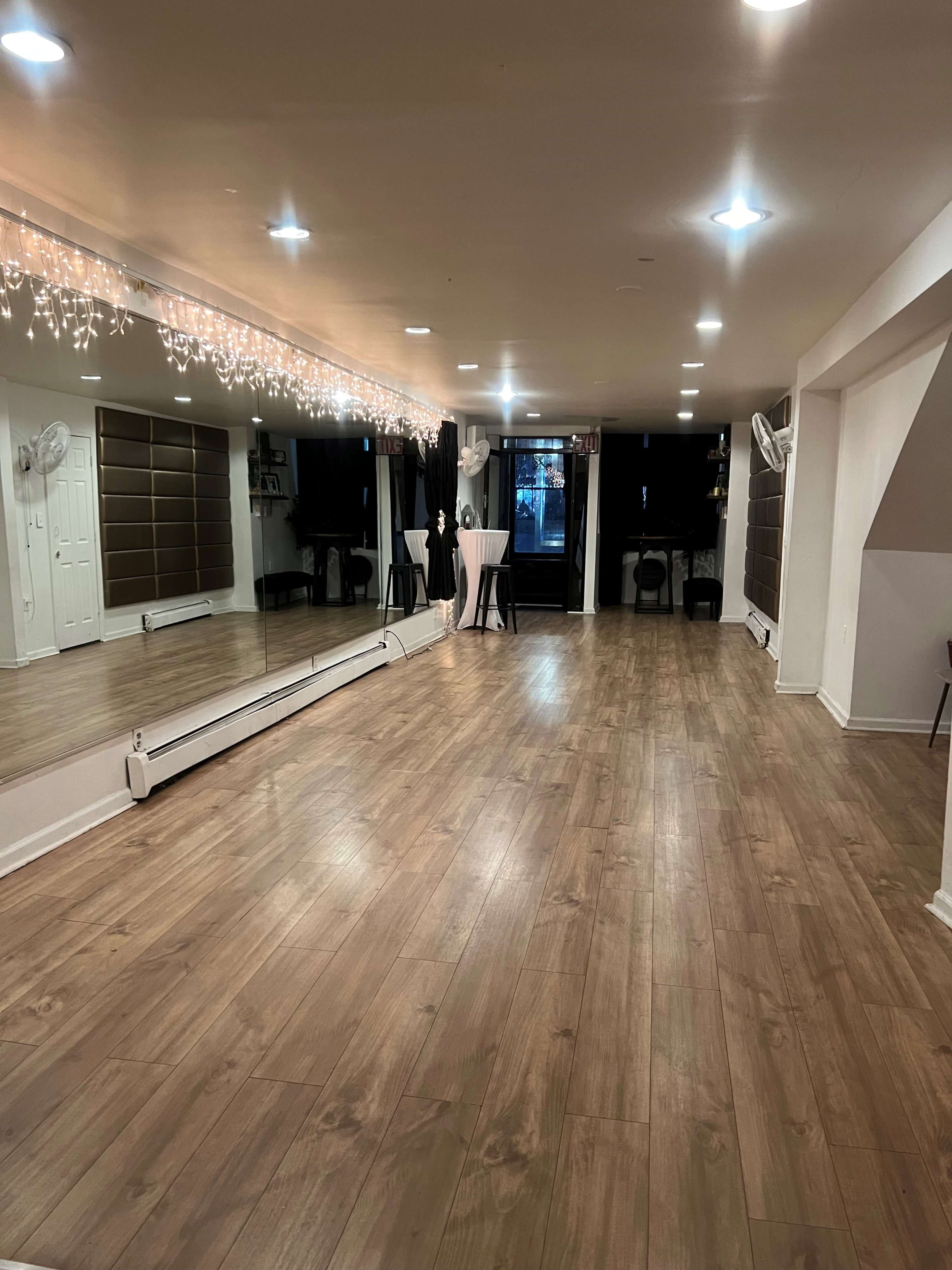 A spacious room with wooden flooring, large mirrors on one wall, and soft lighting from a ceiling decorated with string lights.