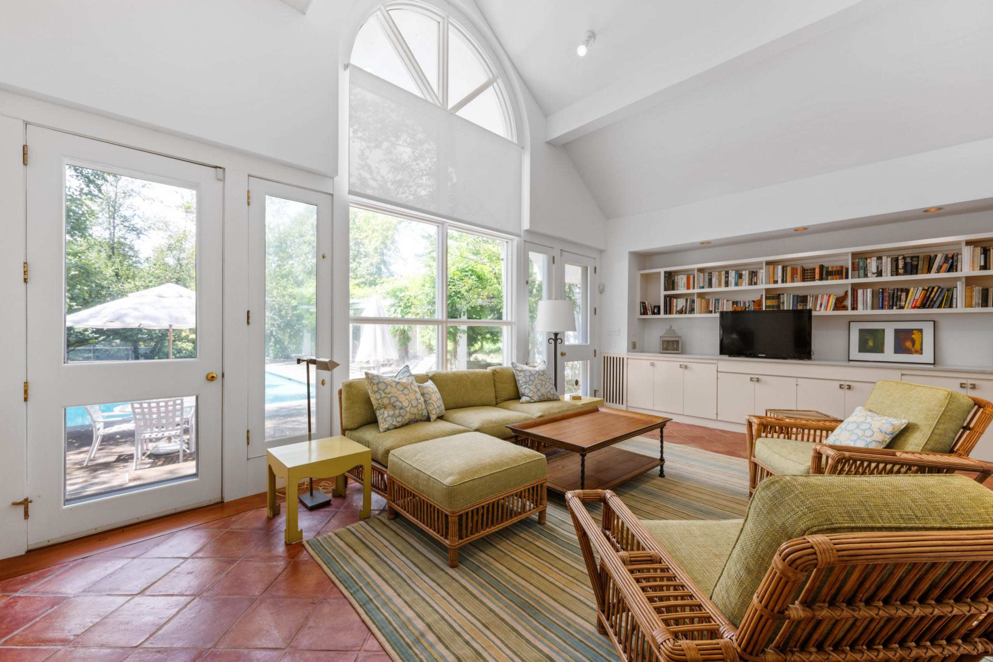 The image shows a bright living room with large windows, wooden furniture, and access to an outdoor pool area.