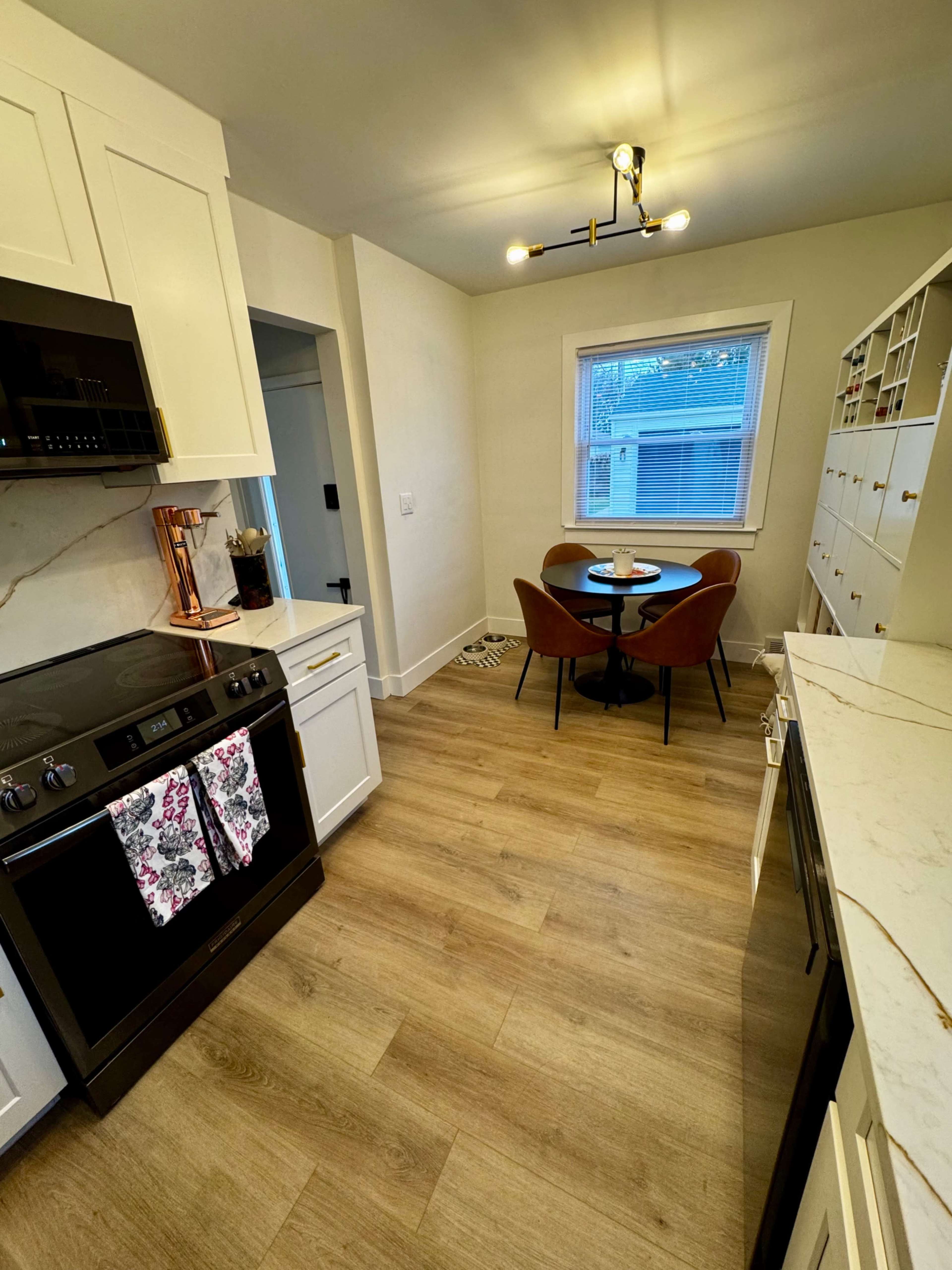 The image shows a modern kitchen featuring white cabinetry, a black oven, a round dining table with brown chairs, and a window allowing natural light.