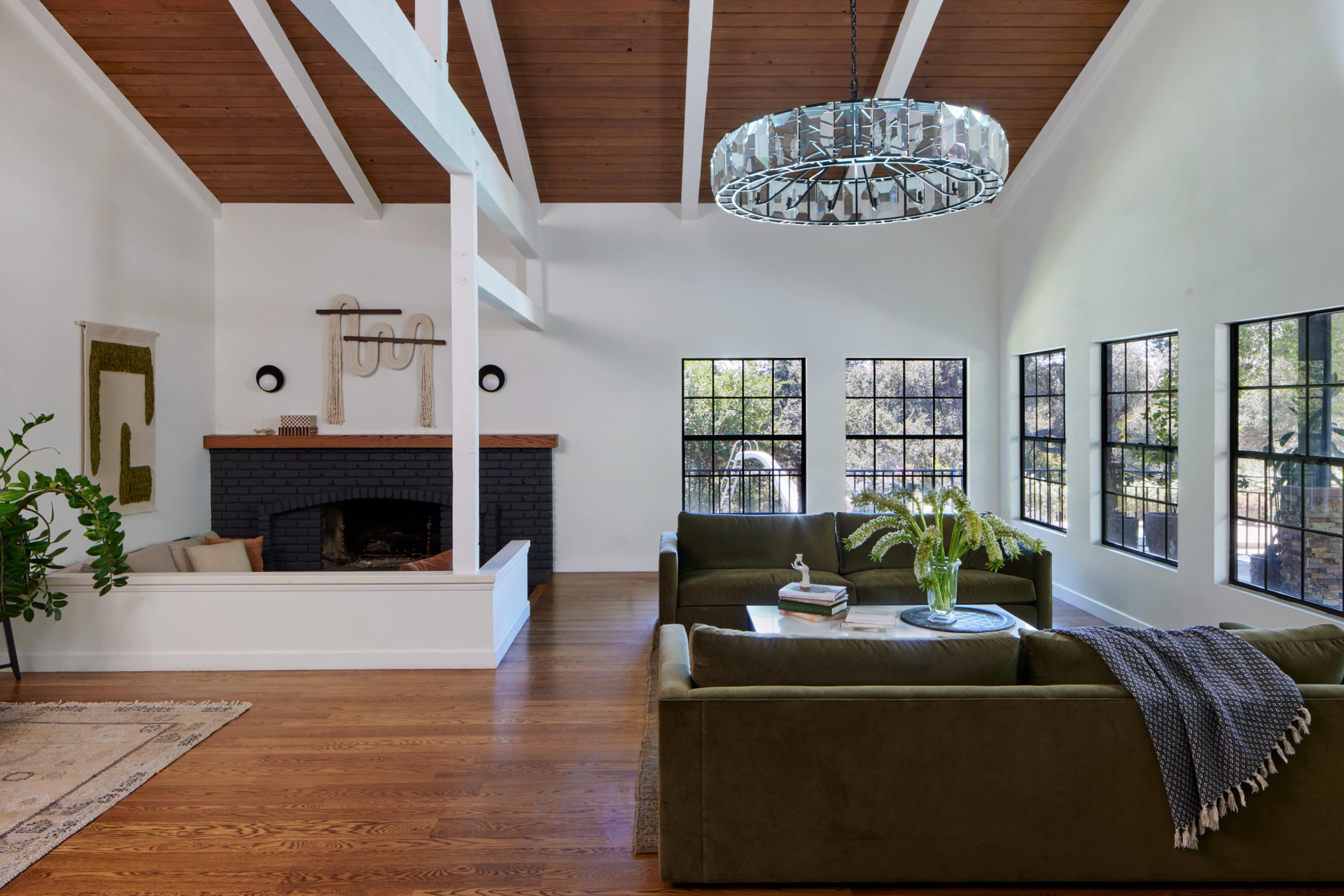 The image shows a modern living room with a high ceiling, large windows, a black fireplace, and green sofas arranged around a coffee table.