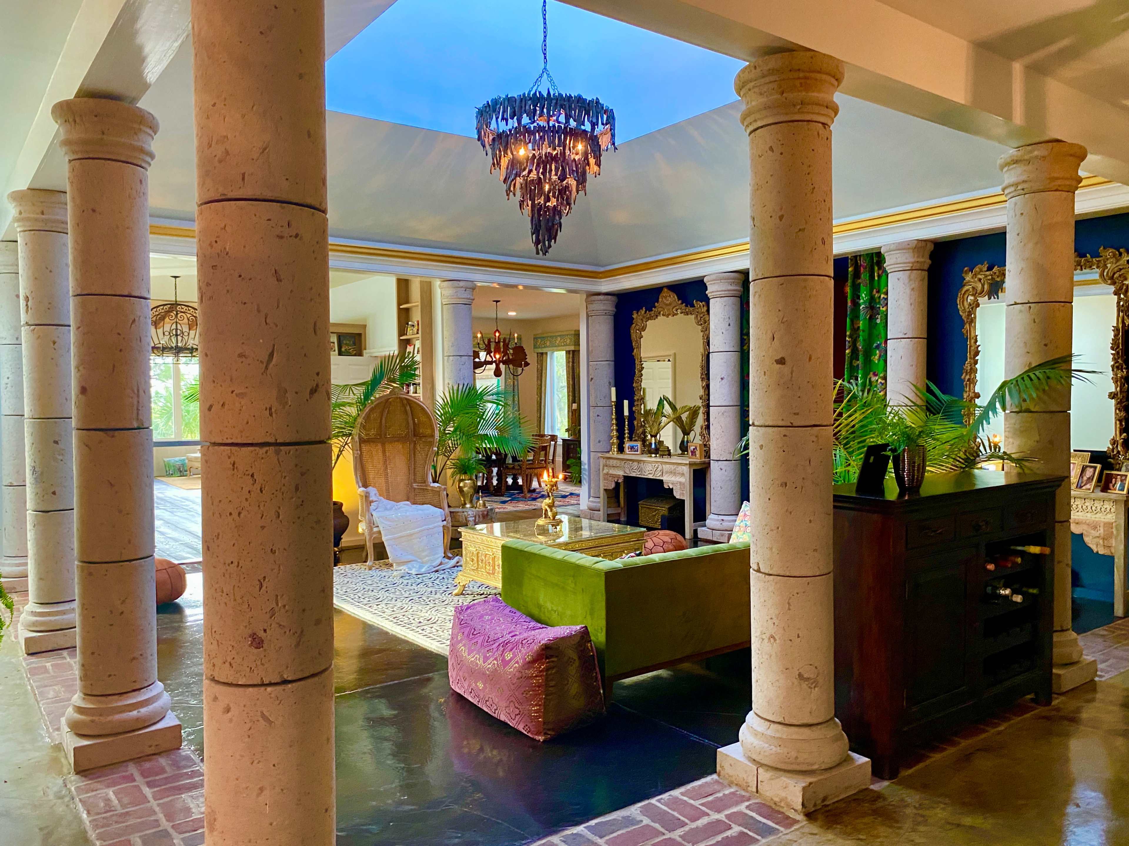 A spacious living area with tall columns, a chandelier, and a mix of decorative furniture and plants.