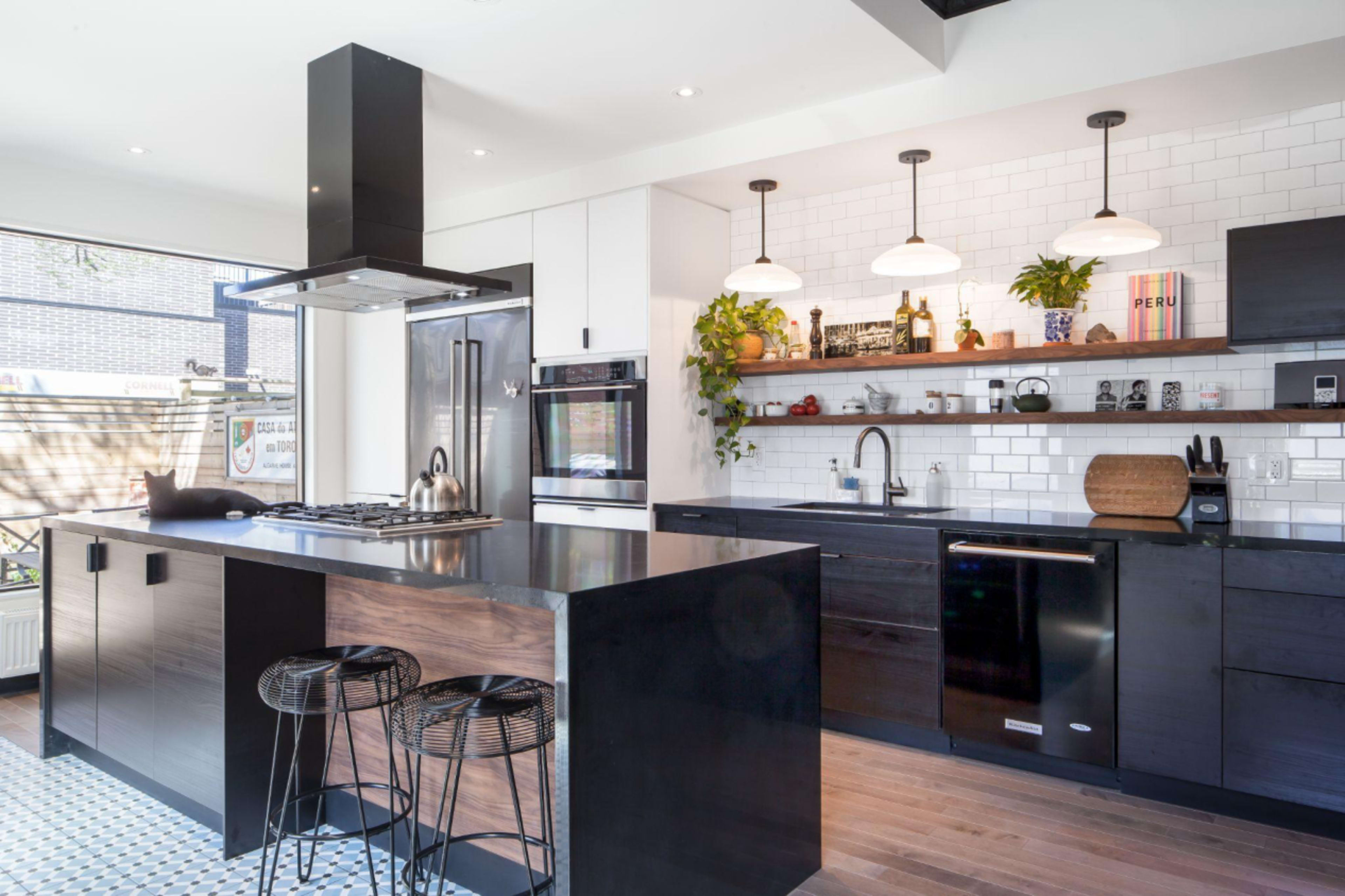 A modern kitchen featuring a large black island, stainless steel appliances, and a wall with open shelves displaying plants and decorative items.