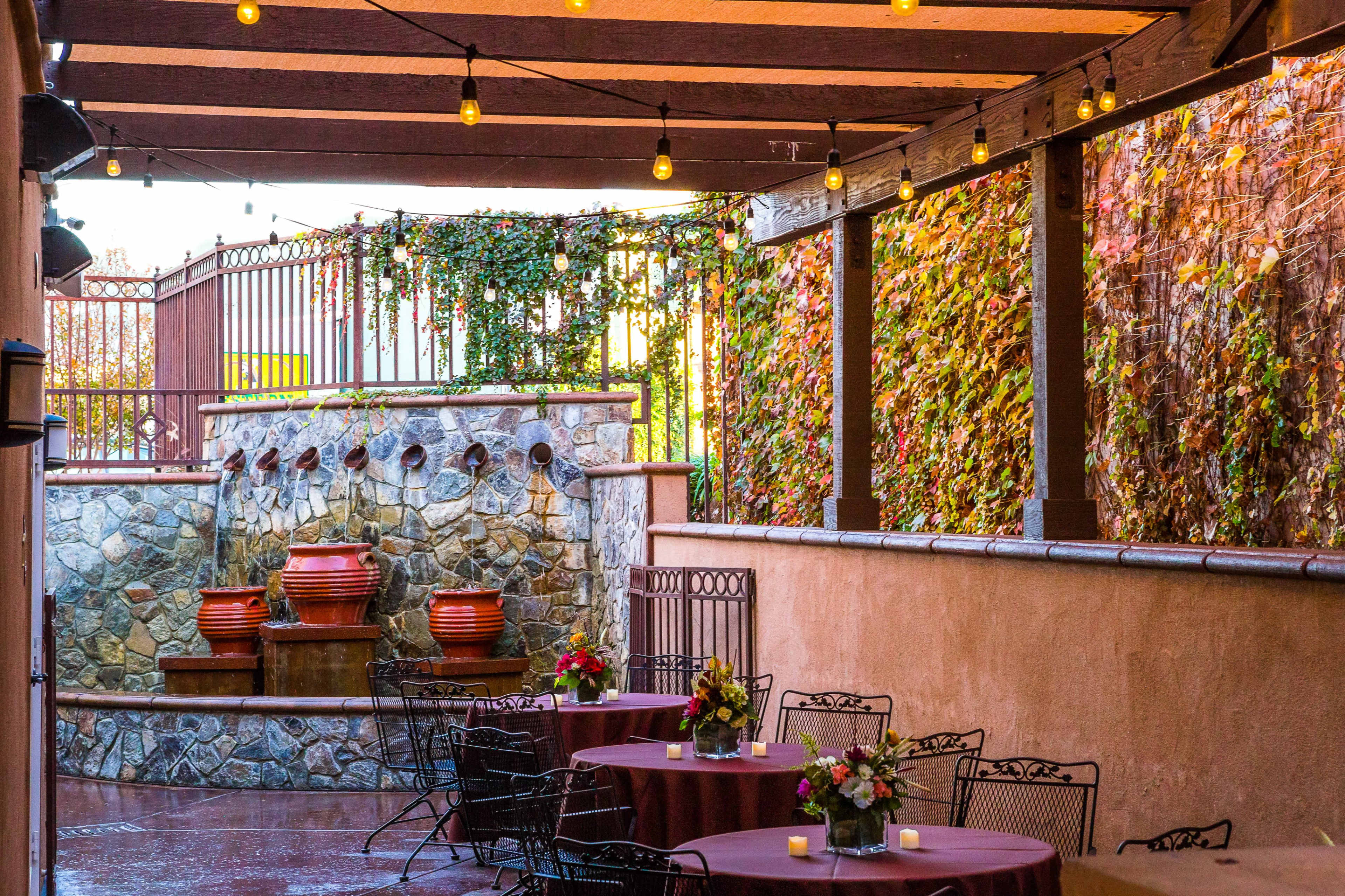 An outdoor dining area with stone walls, decorative planters, and tables set with floral centerpieces under string lights.