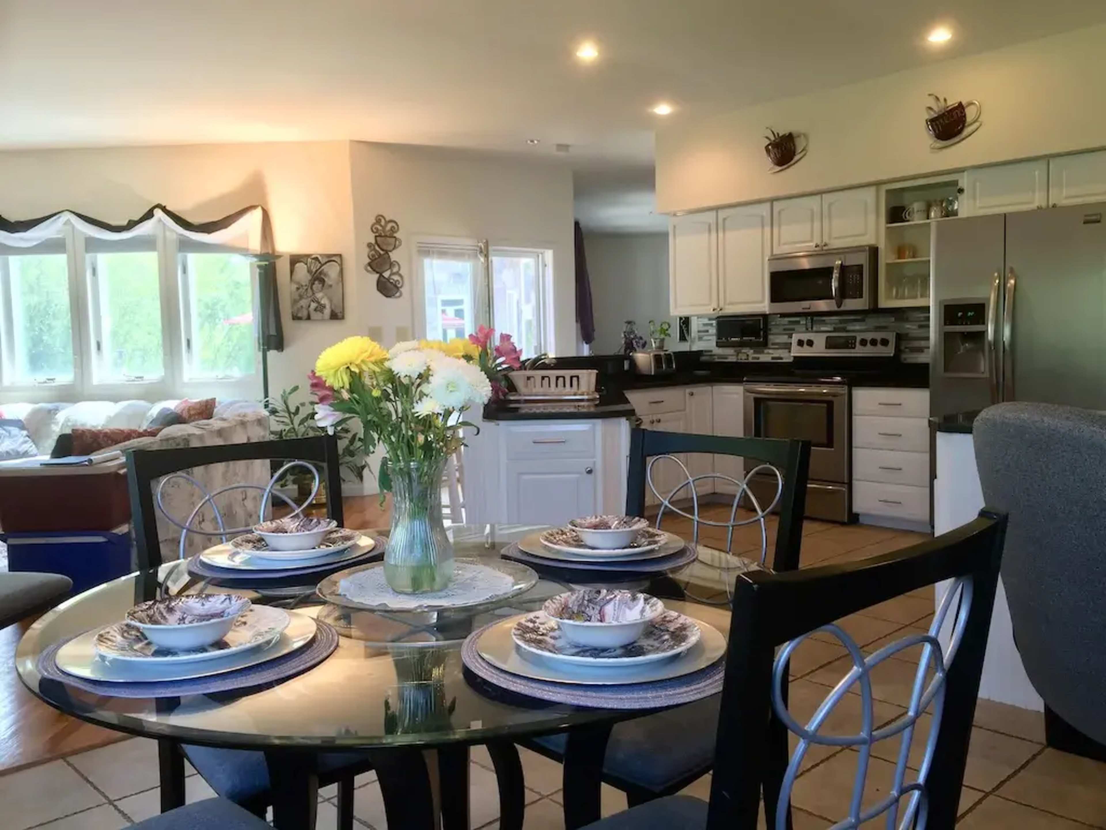 A dining table with floral centerpieces and place settings is set in a modern kitchen area featuring stainless steel appliances and ample natural light.