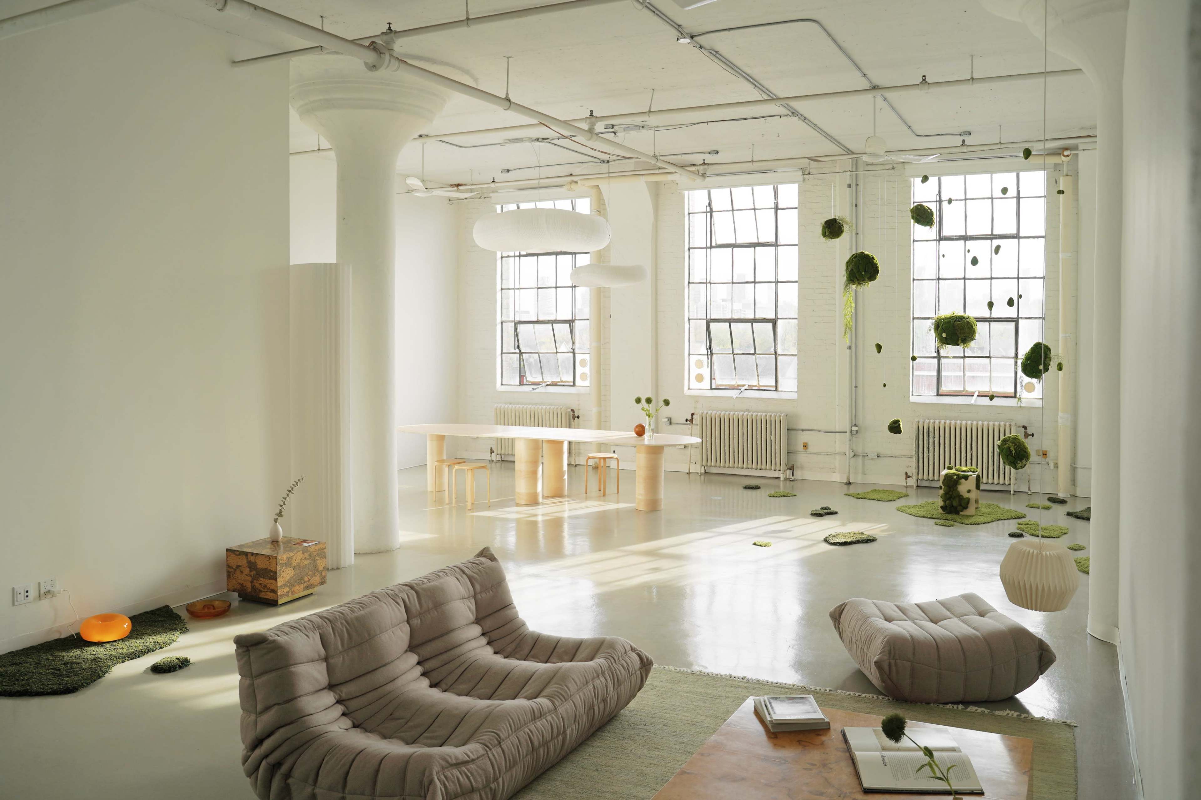 The image shows a minimalist interior space with large windows, featuring light-colored walls, various seating arrangements, and hanging green decor.