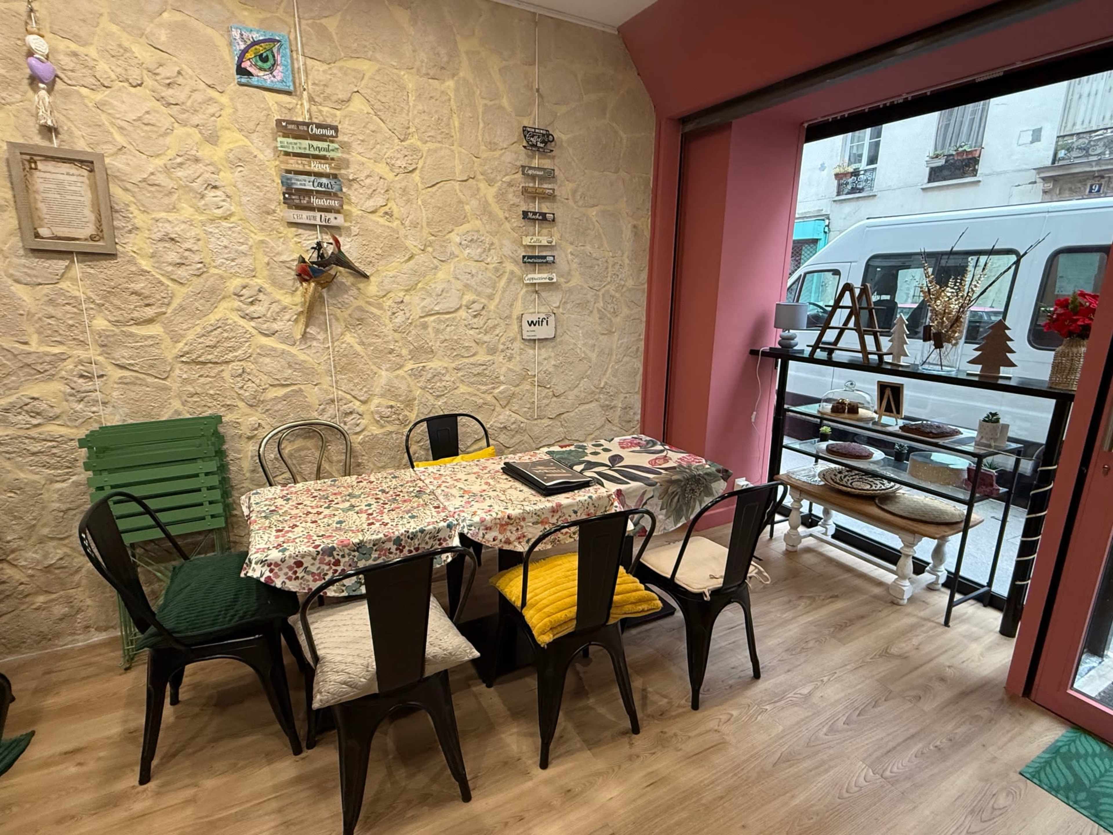 The image shows a dining area with a long table surrounded by black metal chairs, decorated with a floral tablecloth, and a view of the street through a large window.