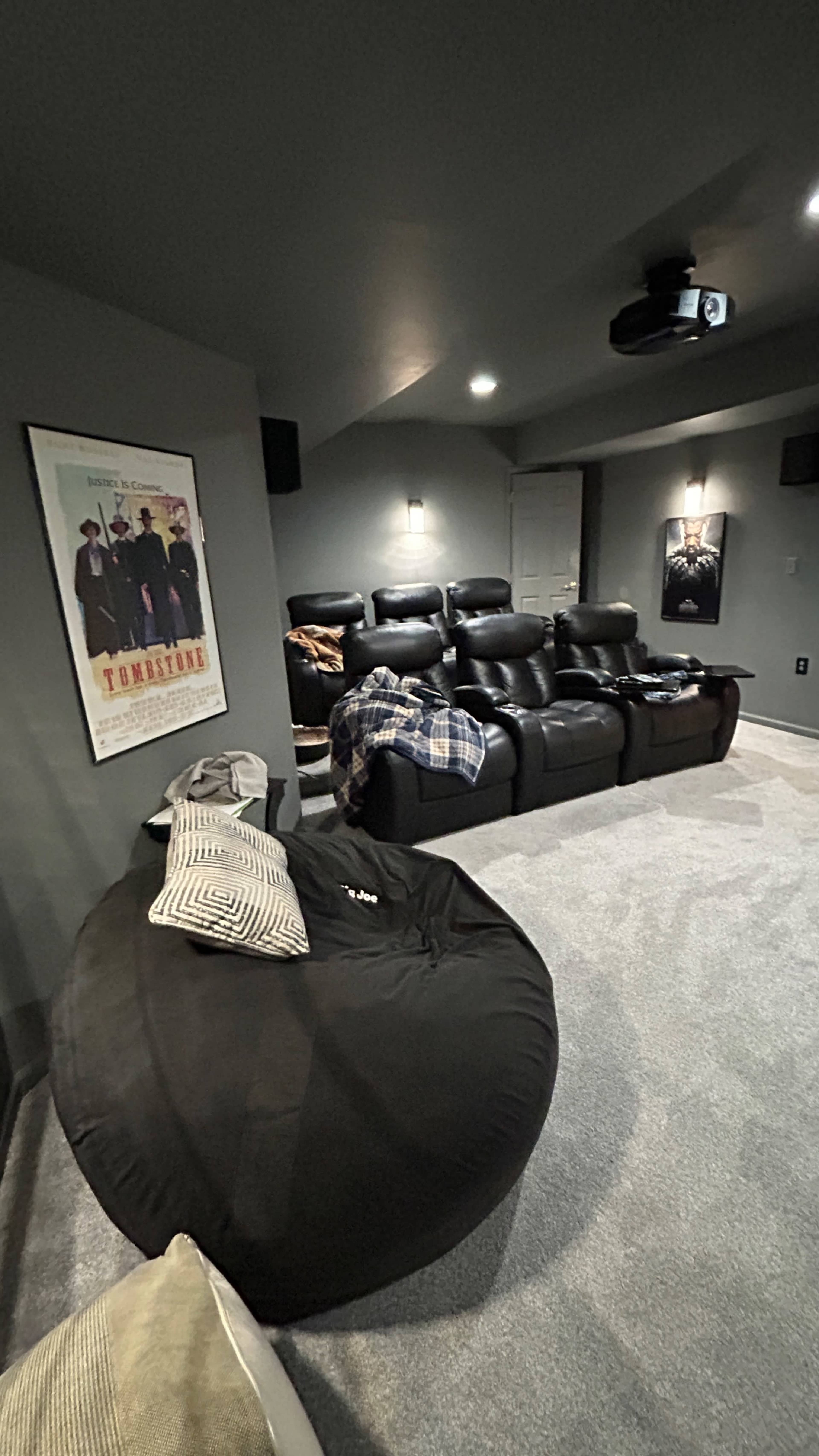 The image shows a home theater room with black reclining chairs, a projector on the ceiling, and a large bean bag chair on the floor next to a wall featuring a movie poster.