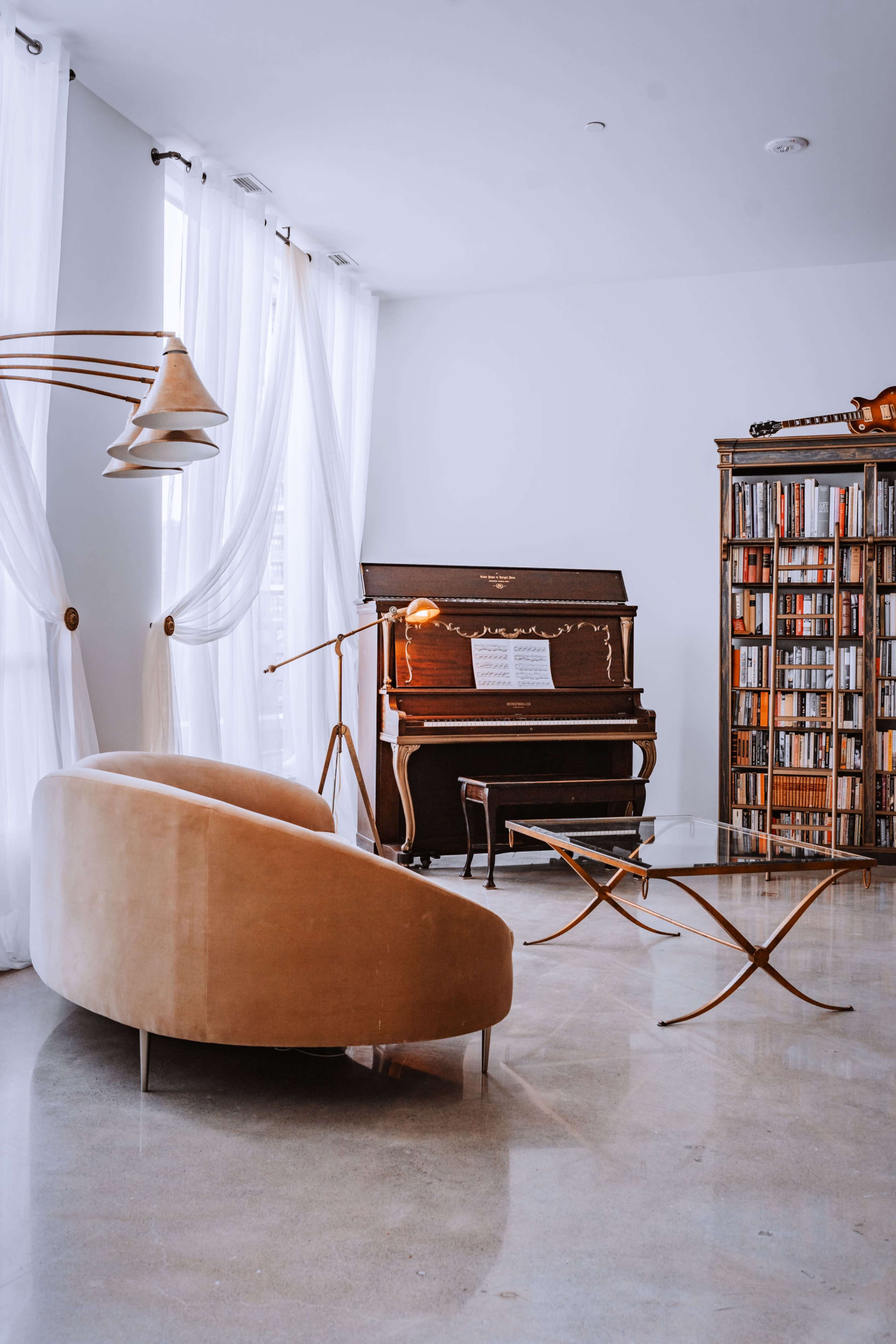 The image shows a modern living room featuring a curved beige sofa, a piano against the wall, a bookshelf filled with books, and large windows with flowing white curtains.