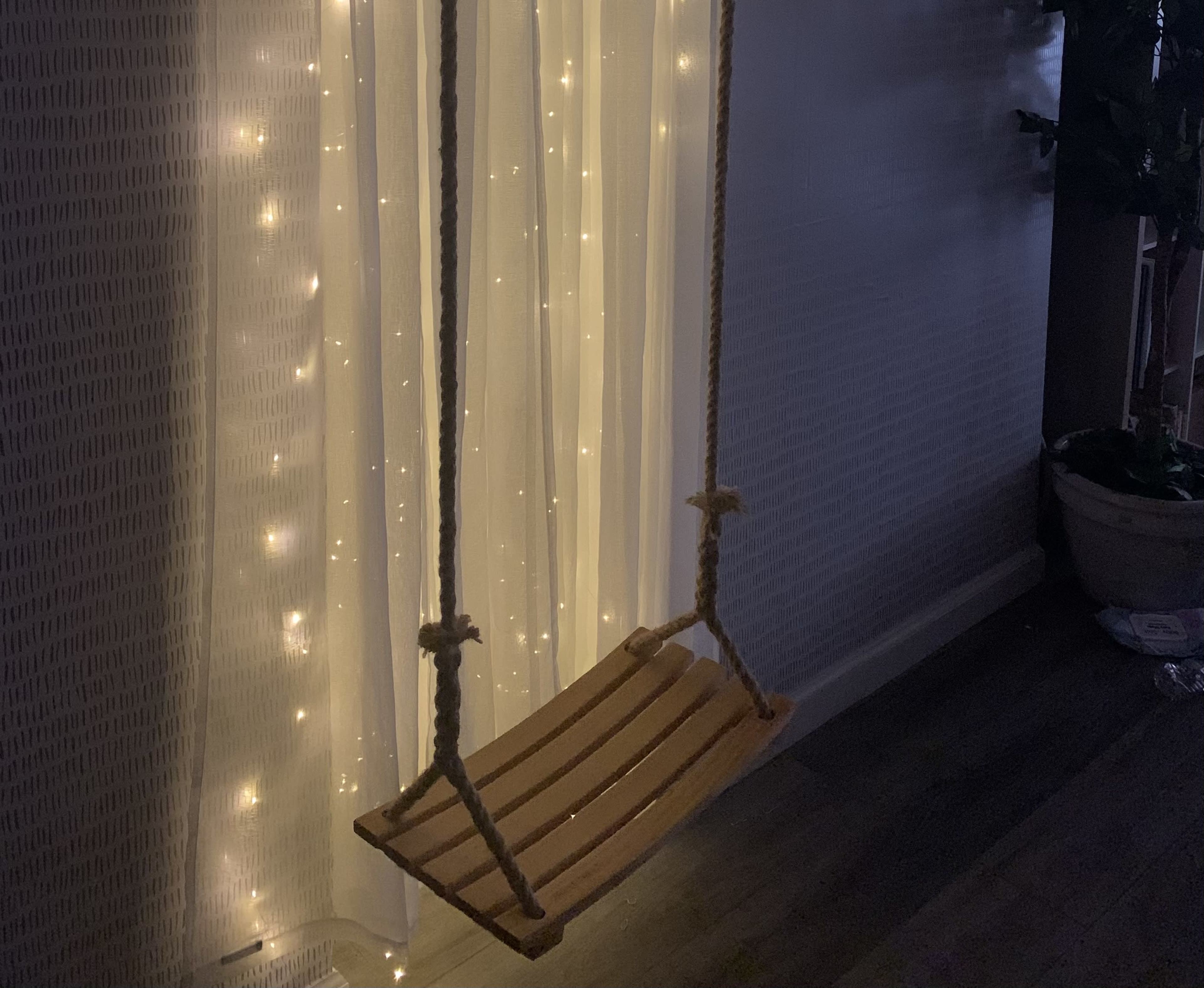 A wooden swing hangs from the ceiling in front of a wall adorned with string lights.