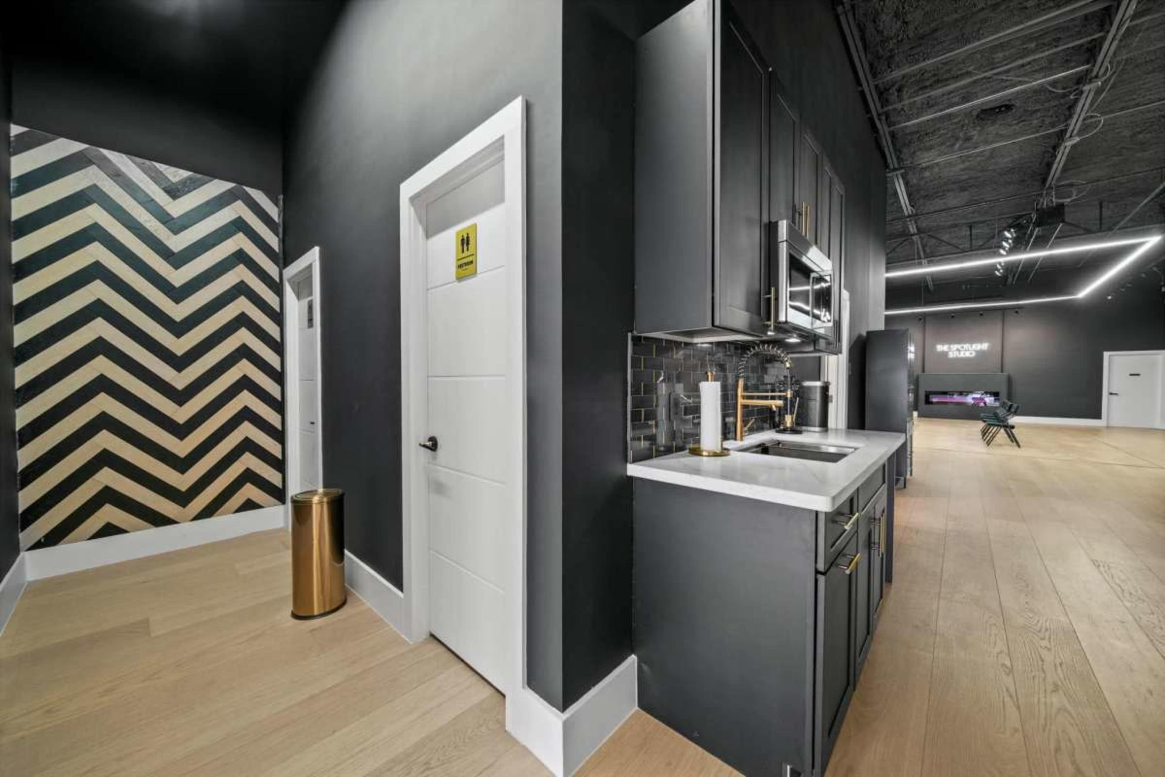 The image shows a modern kitchen area with dark cabinetry, a white countertop, and a stylish geometric accent wall, leading to a hallway with a door and a warning sign.