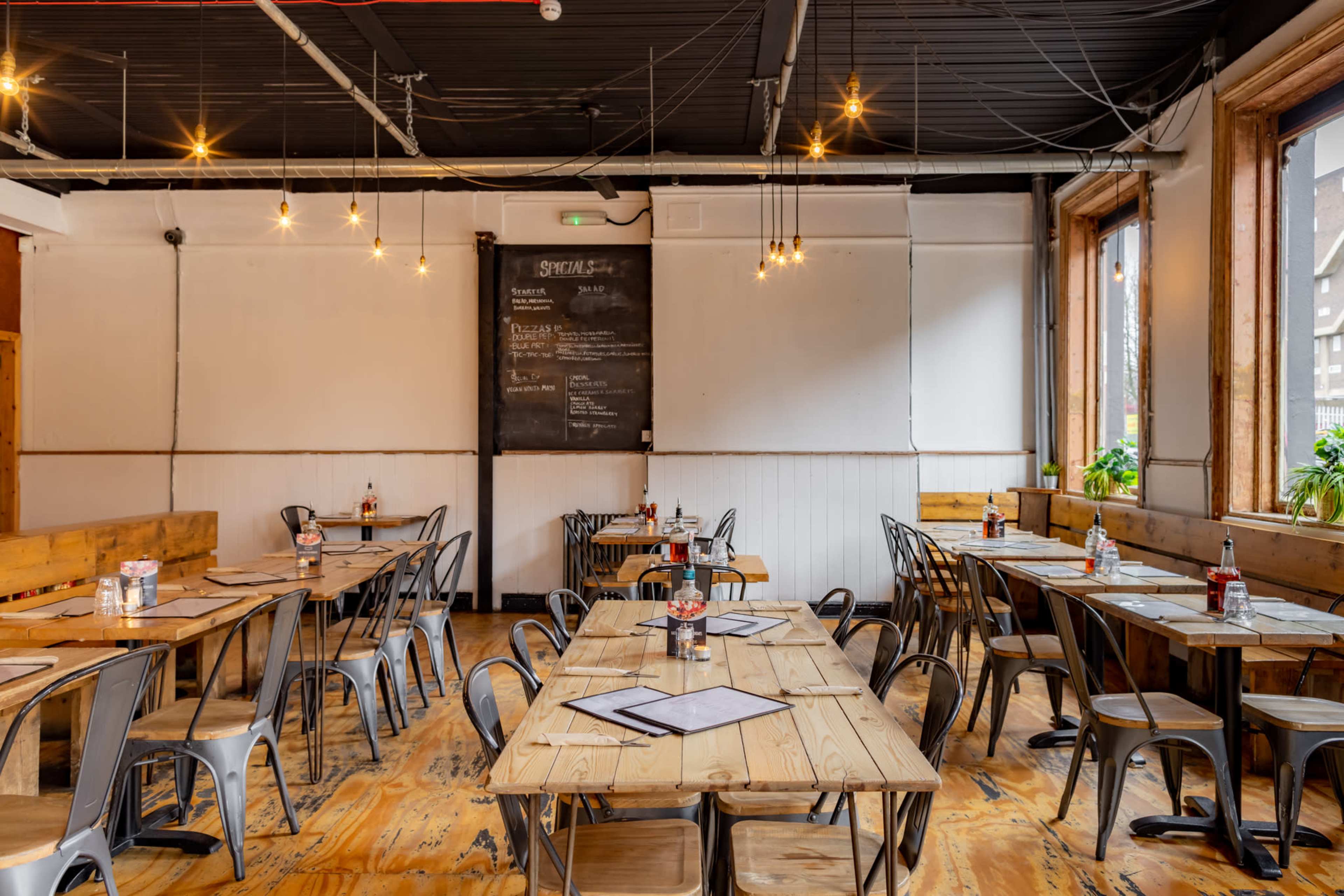 The image shows a spacious restaurant with wooden tables, metallic chairs, and large windows, illuminated by hanging light fixtures.