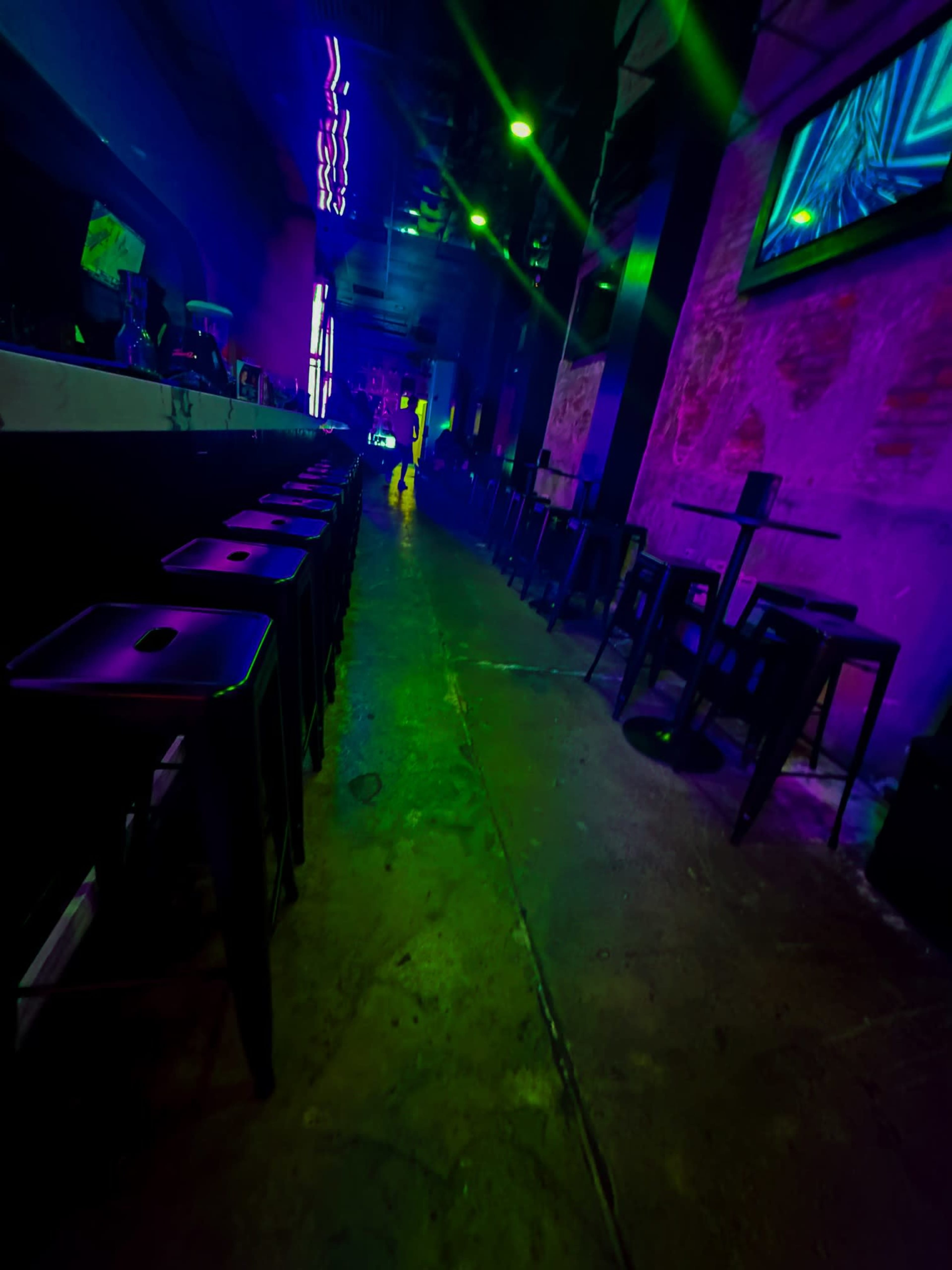 The image shows a dimly lit bar or club interior with a long bar counter on one side and high tables and stools arranged on the floor.