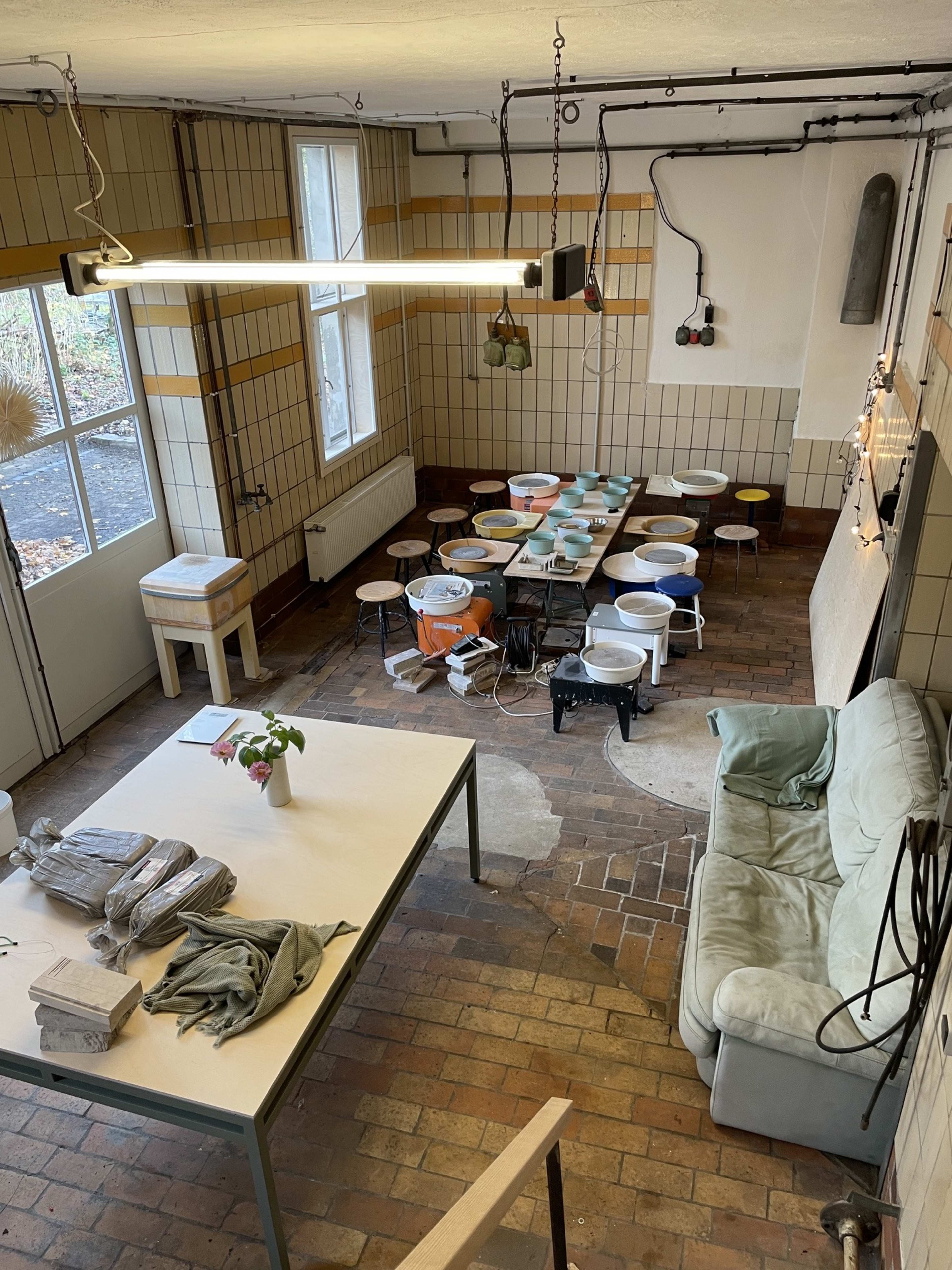 The image shows a spacious room with tiled walls, a seating area with a light green couch, a central table cluttered with materials, and an array of colorful stools arranged in rows.