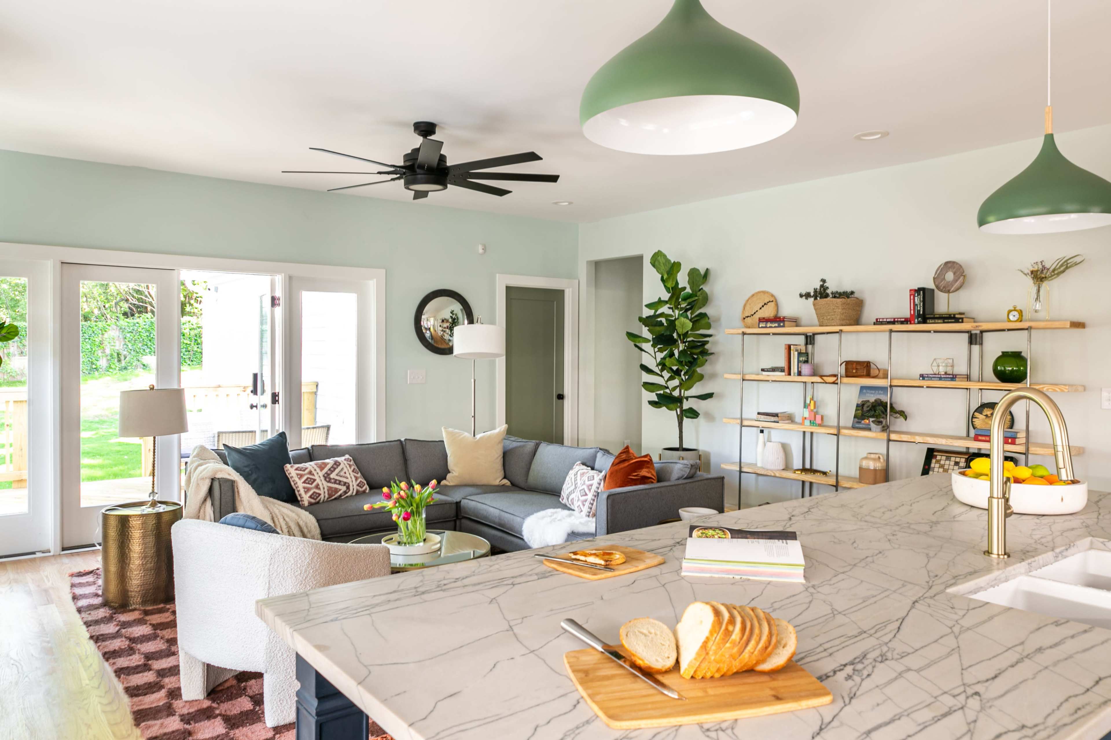 A modern open-concept living area features a gray sectional sofa, a marble countertop with sliced bread, and shelves displaying decorative items.