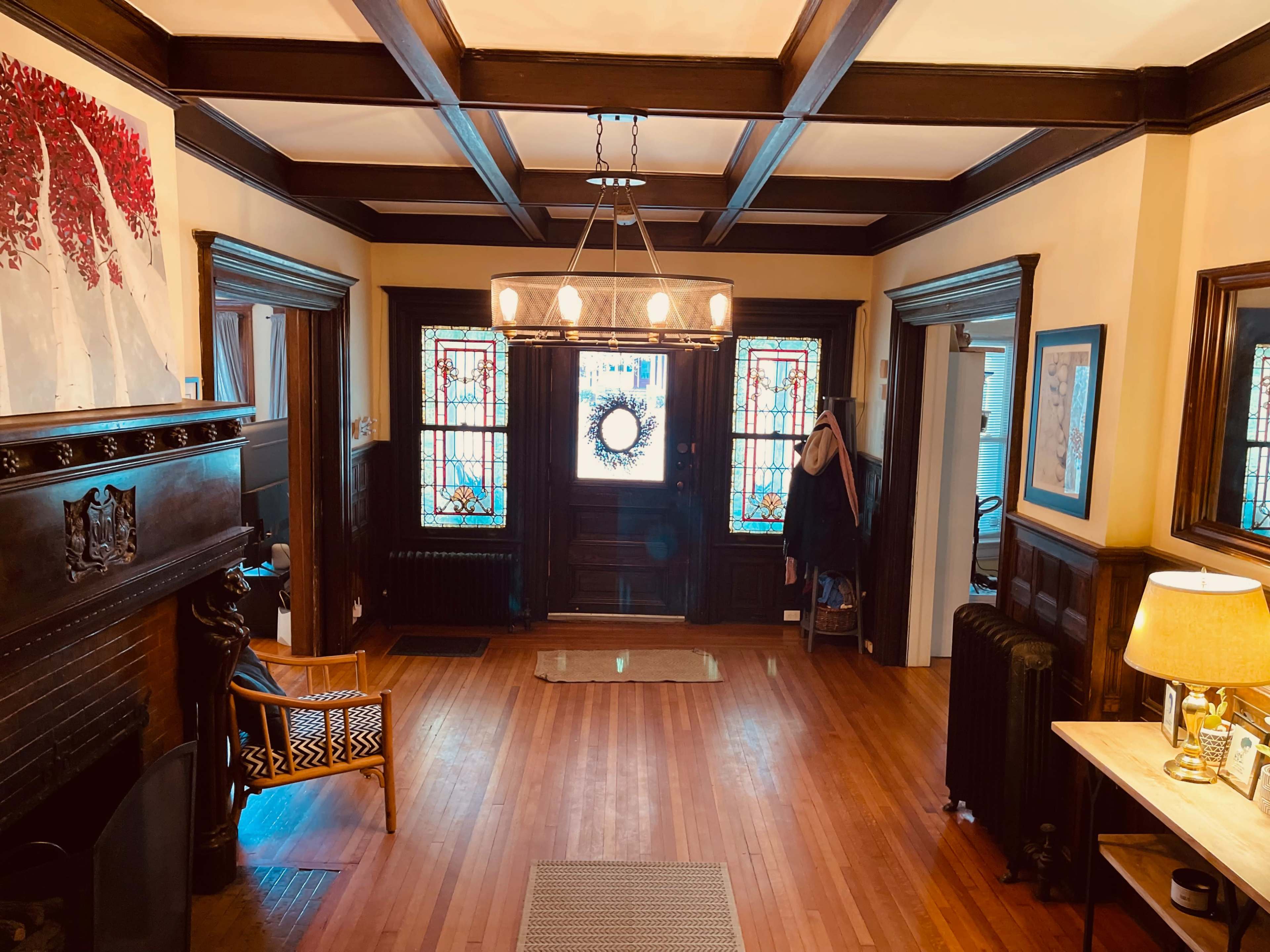 An entrance hall with wooden beams on the ceiling, stained glass windows on either side of a central door, and a decorative fireplace.