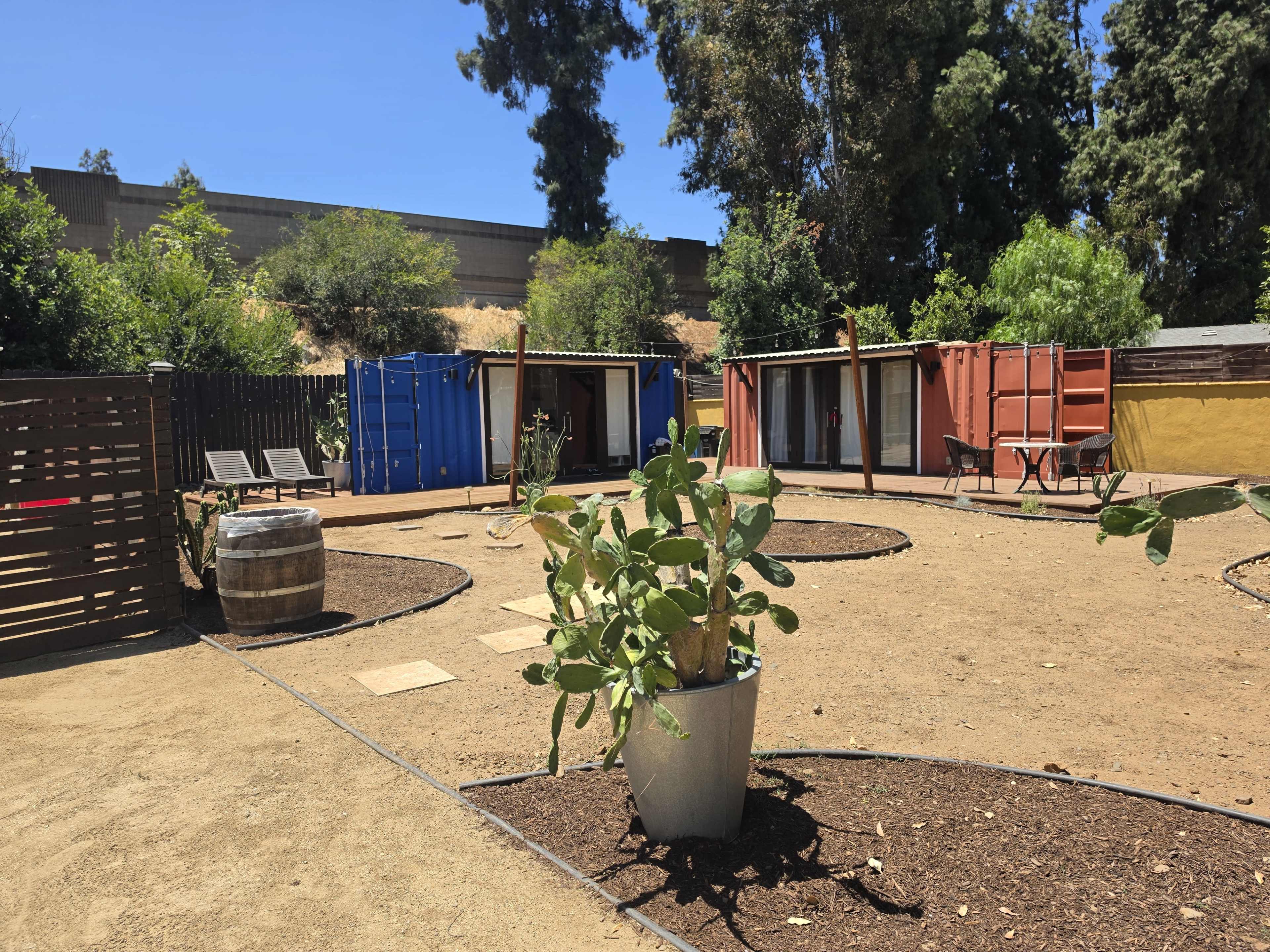 The image shows a landscaped outdoor area featuring several shipping containers, a barrel, and cacti, surrounded by gravel and greenery.