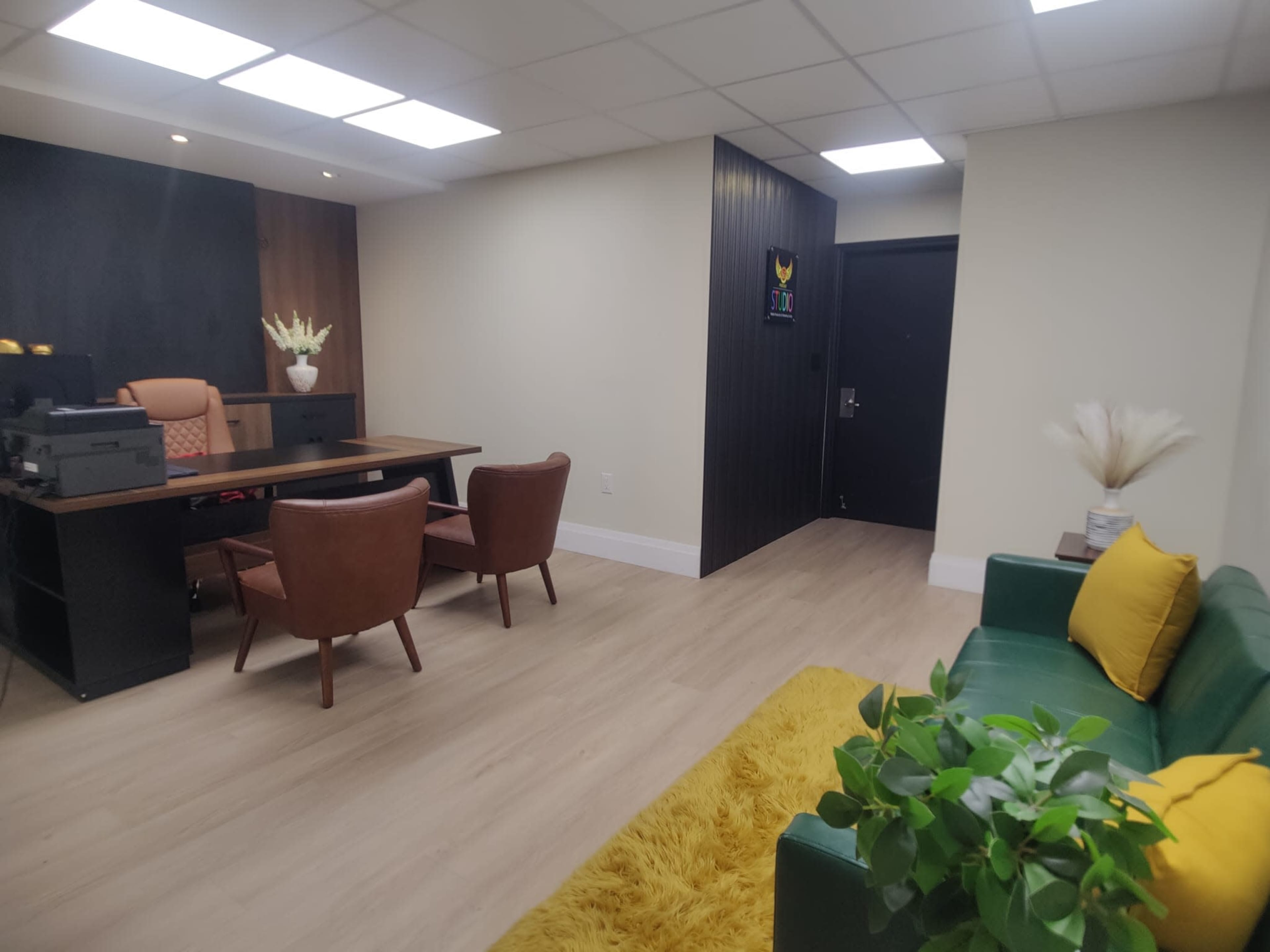 The image shows a modern office space with a wooden desk and two brown chairs, a green couch, and a door leading to another room.