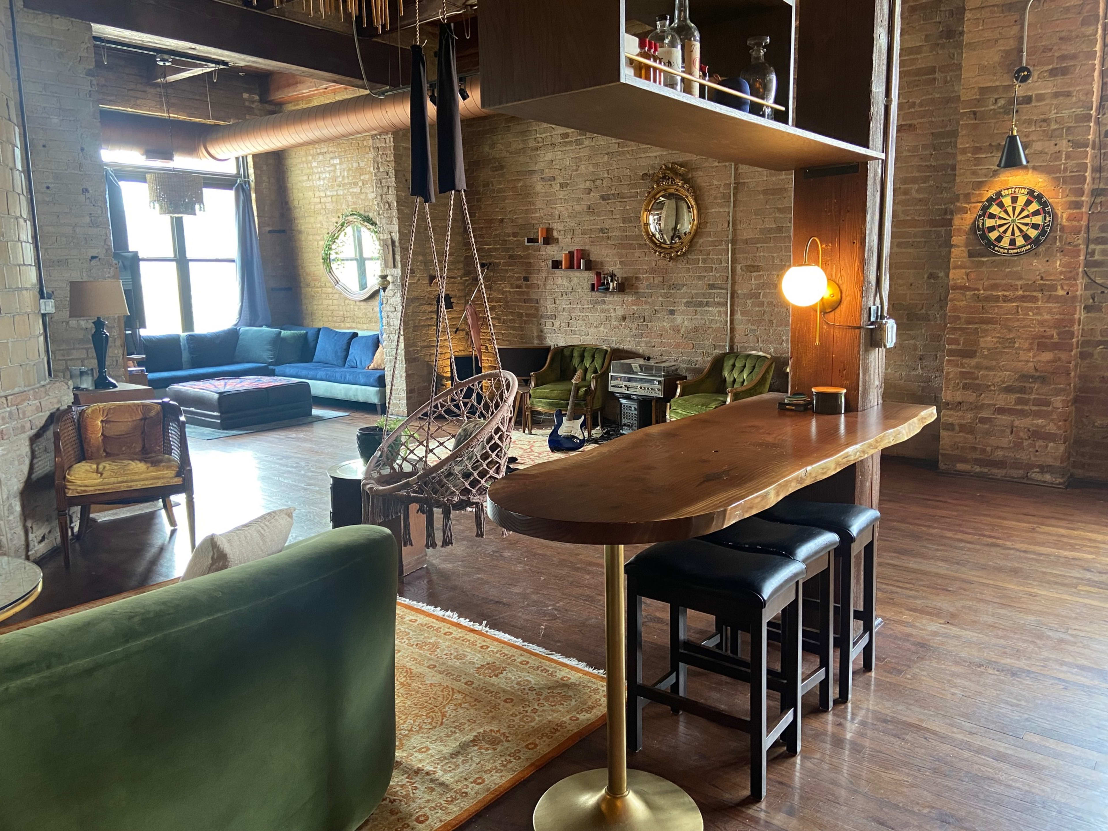 A spacious, modern living area with a mix of seating options, including a hanging chair and a bar counter, set against exposed brick walls.