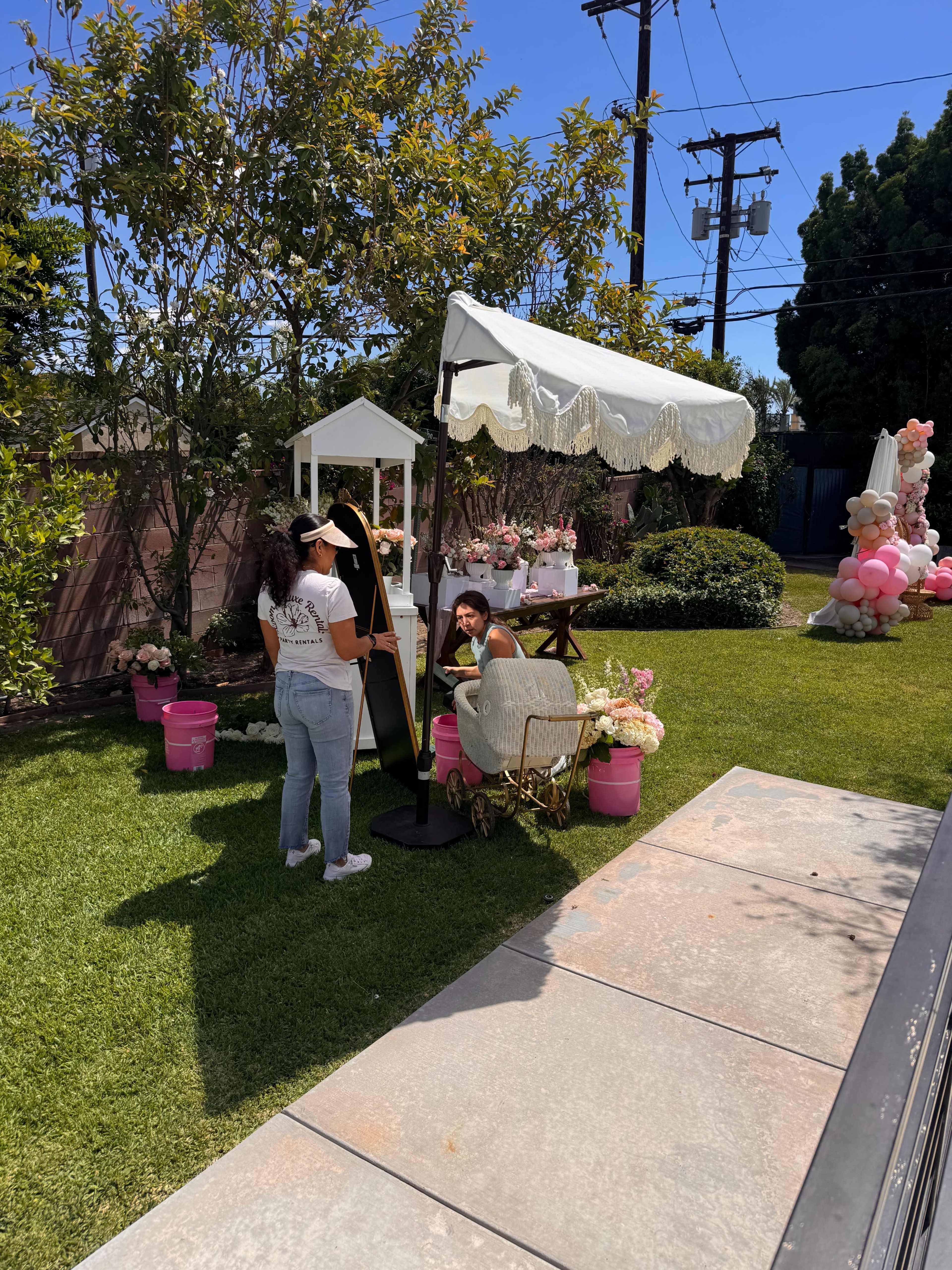 Two women interact with a large mirror setup in a garden adorned with decorative flowers and balloons, while a canopy provides shade.