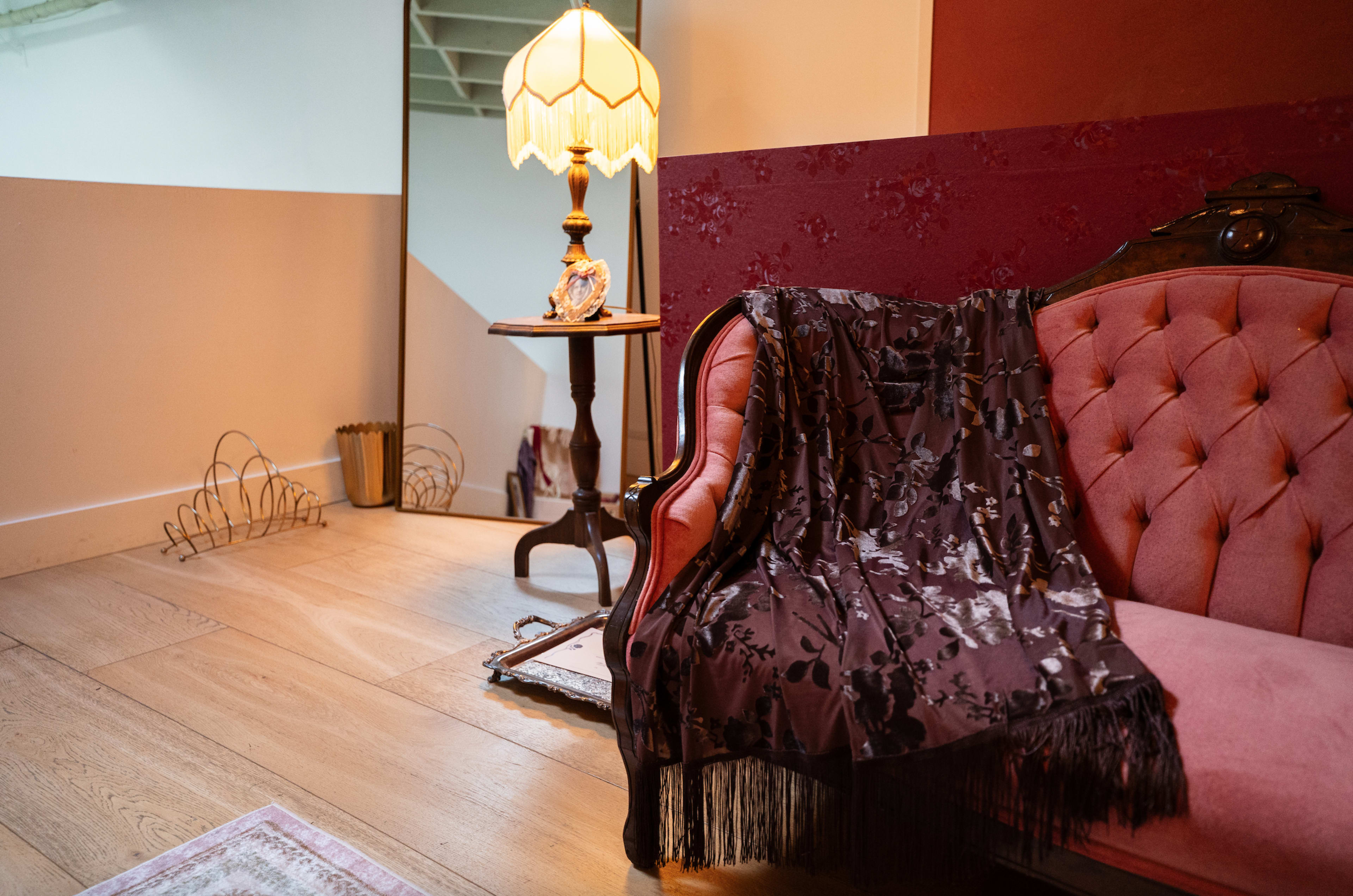 A pink upholstered sofa with a fringed throw is placed next to a lamp and a mirror in a room with wooden flooring.