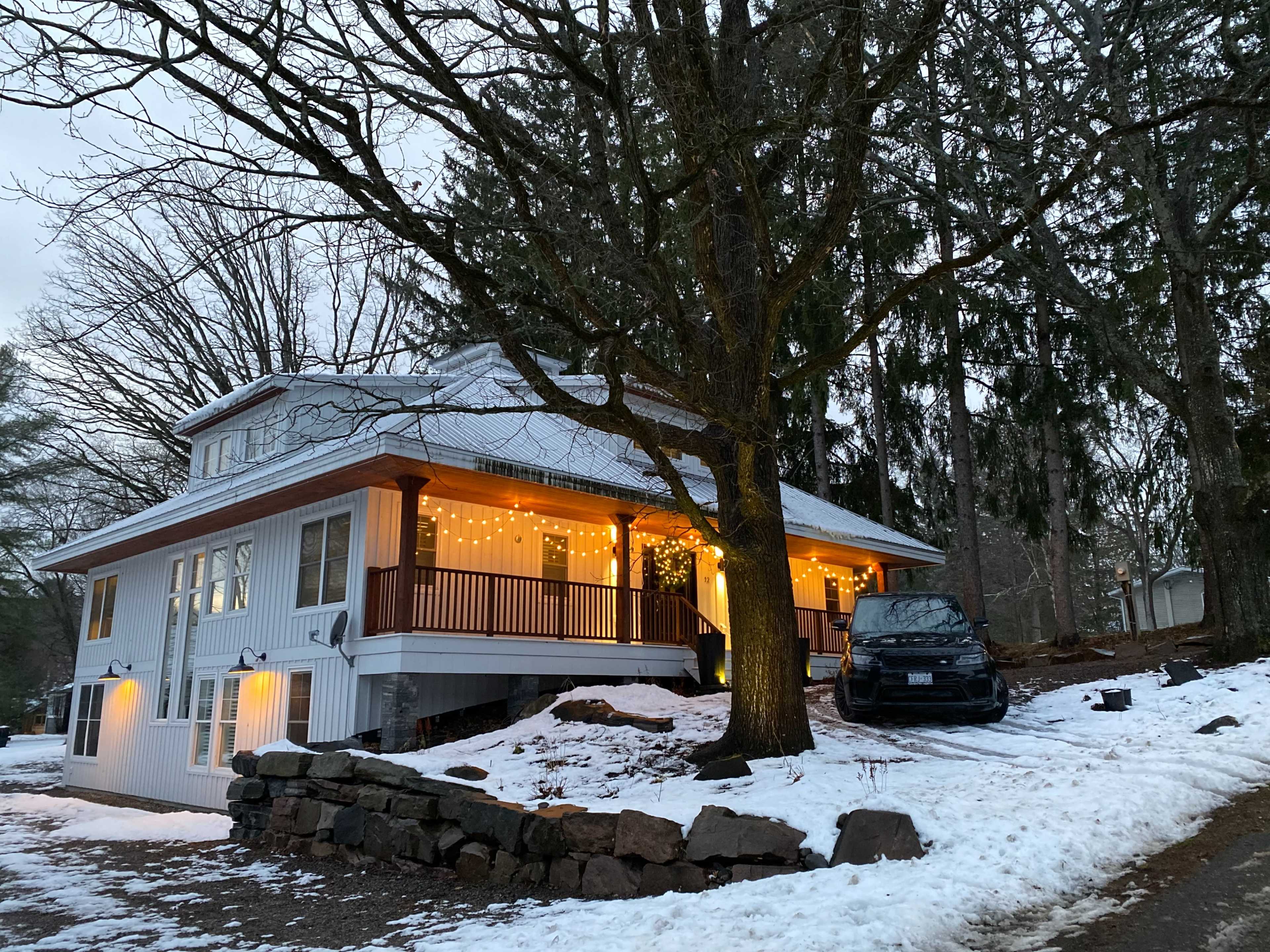 A two-story house with outdoor string lights is situated on a snowy landscape beneath leafless trees.