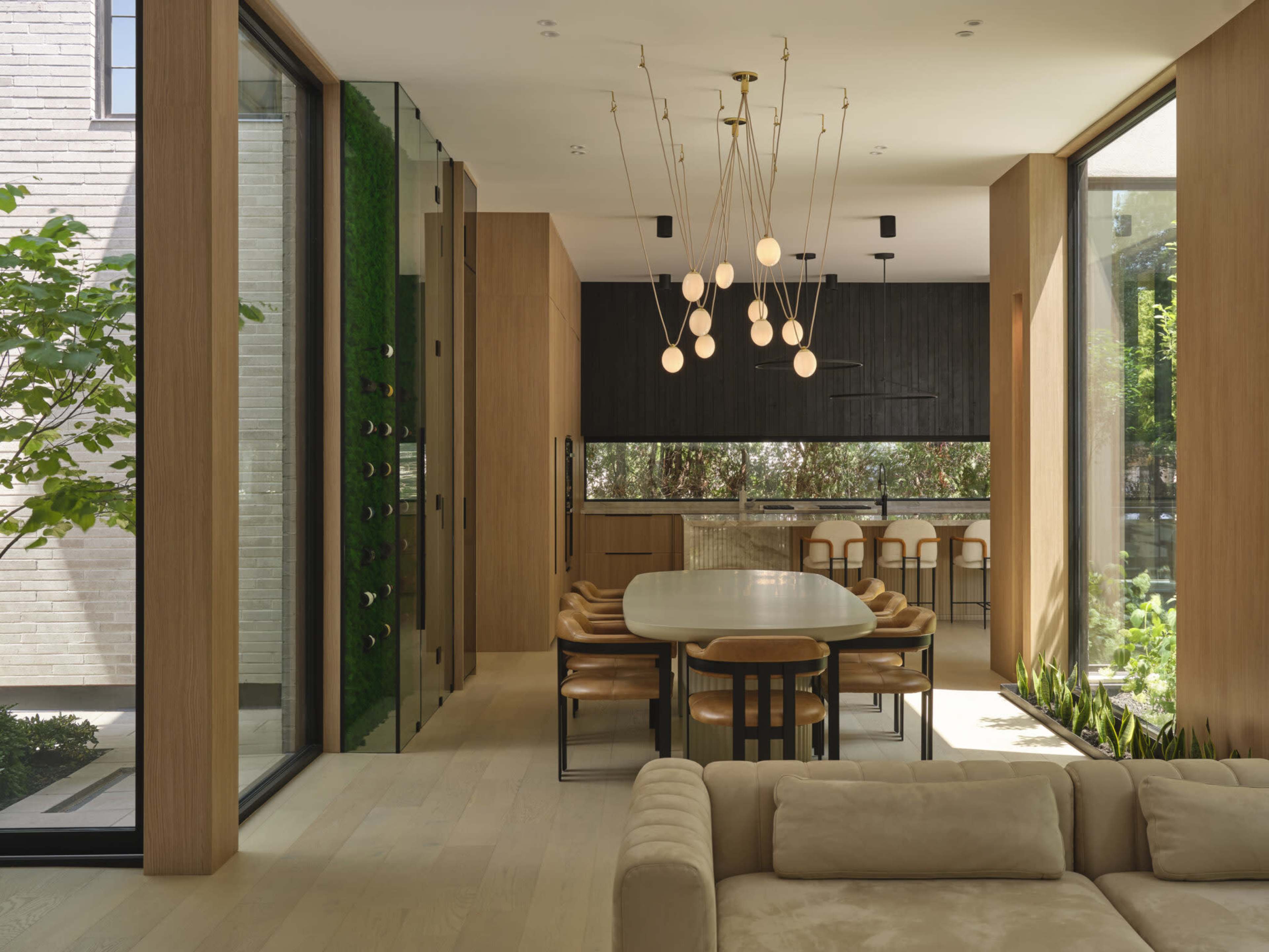 A modern dining area features a large table surrounded by chairs, with large windows providing views of greenery and a kitchen in the background.