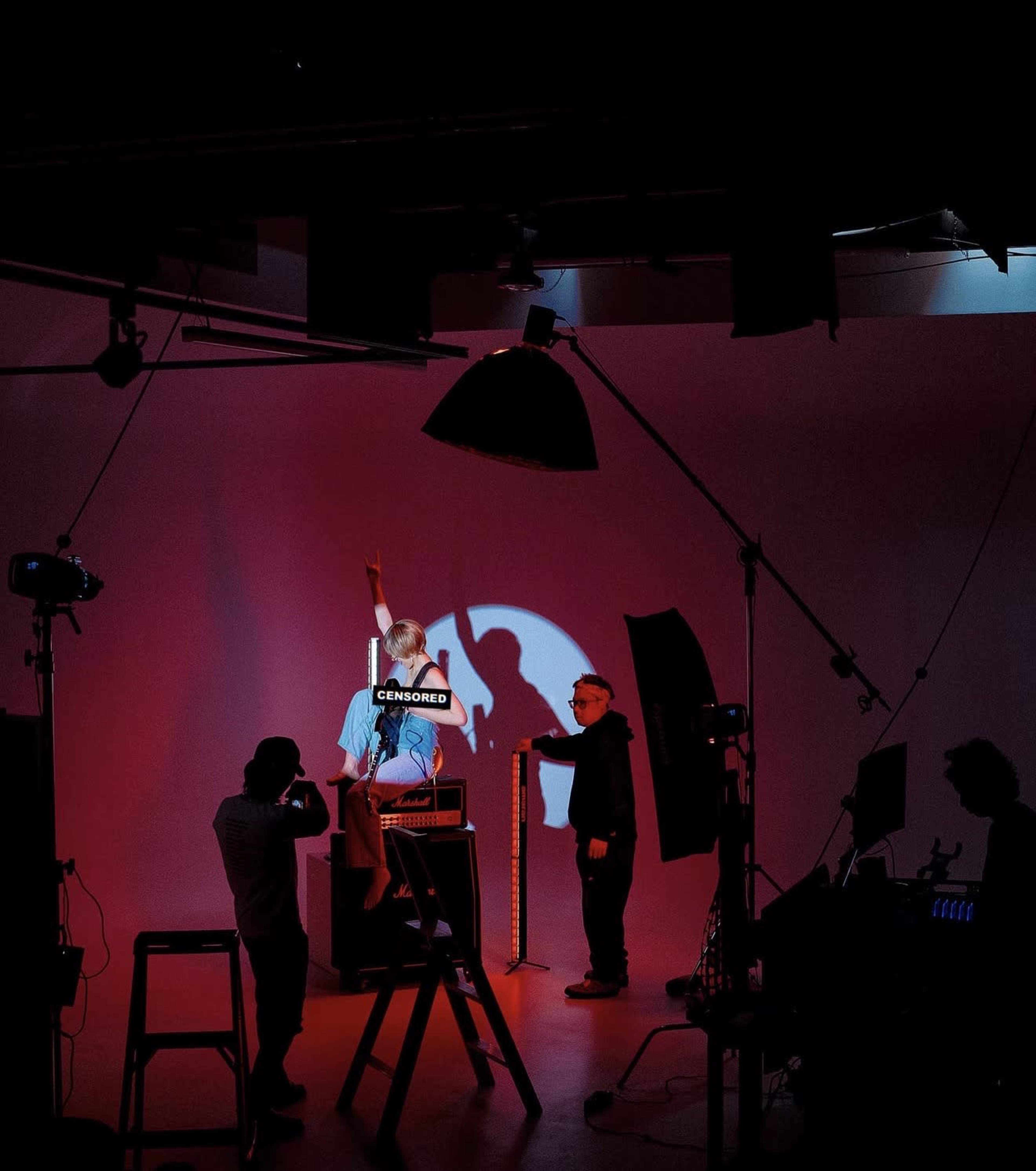 A person stands in front of a backdrop illuminated by colored lights while others adjust equipment and set up the scene.
