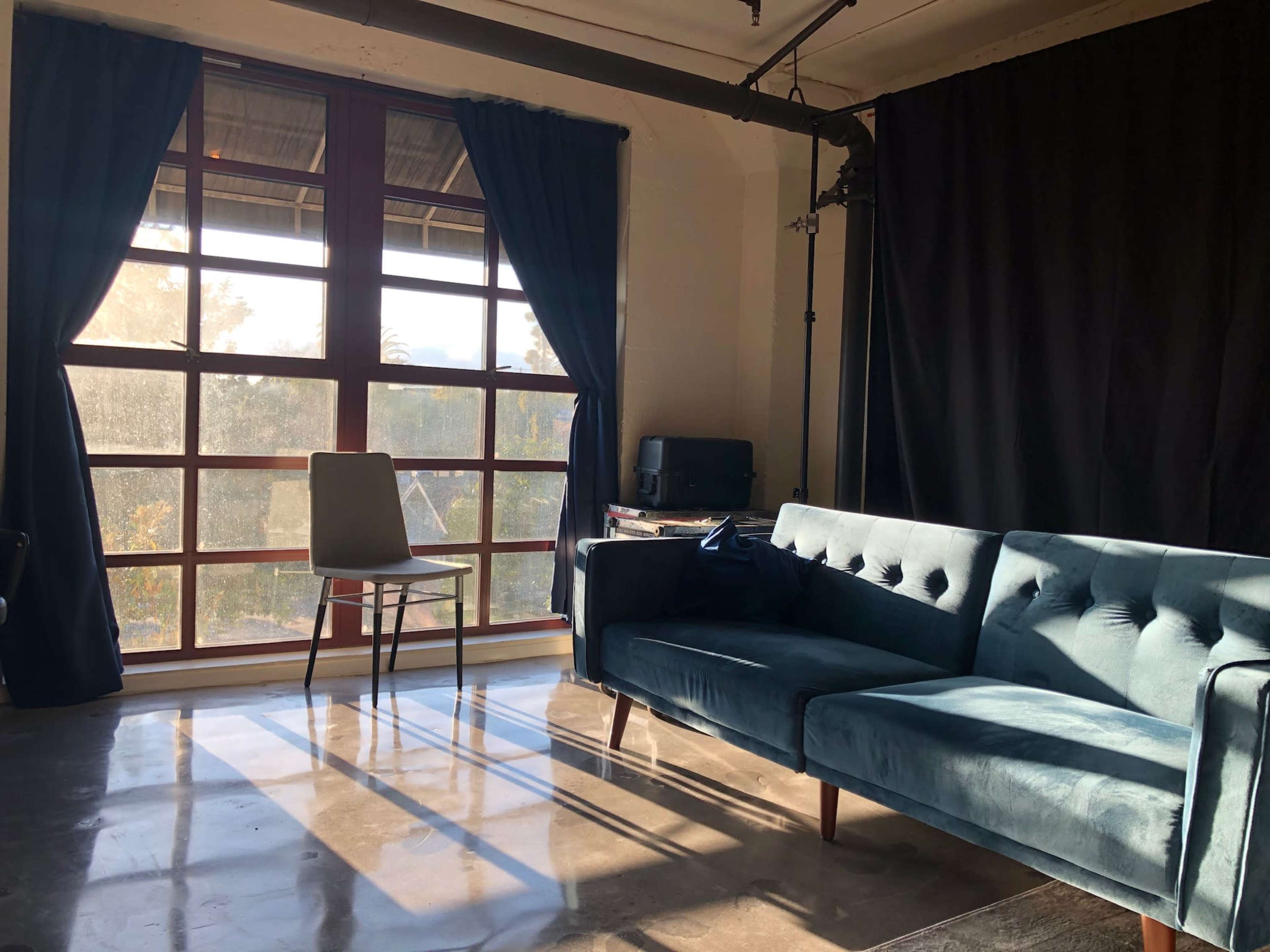 A room features a blue velvet sofa, a chair, and large windows with sunlight casting shadows on the polished floor.