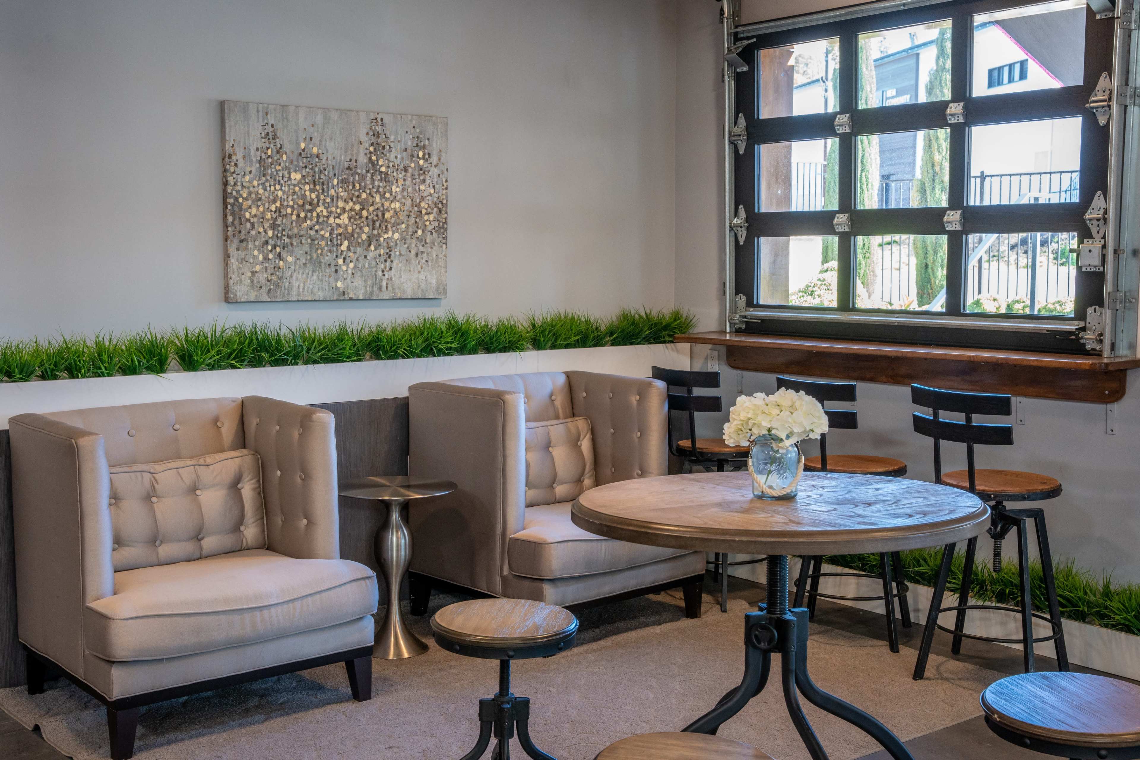 A modern lounge area features two tufted armchairs, a round wooden table, and bar stools, with a decorative painting and potted greenery along the wall.