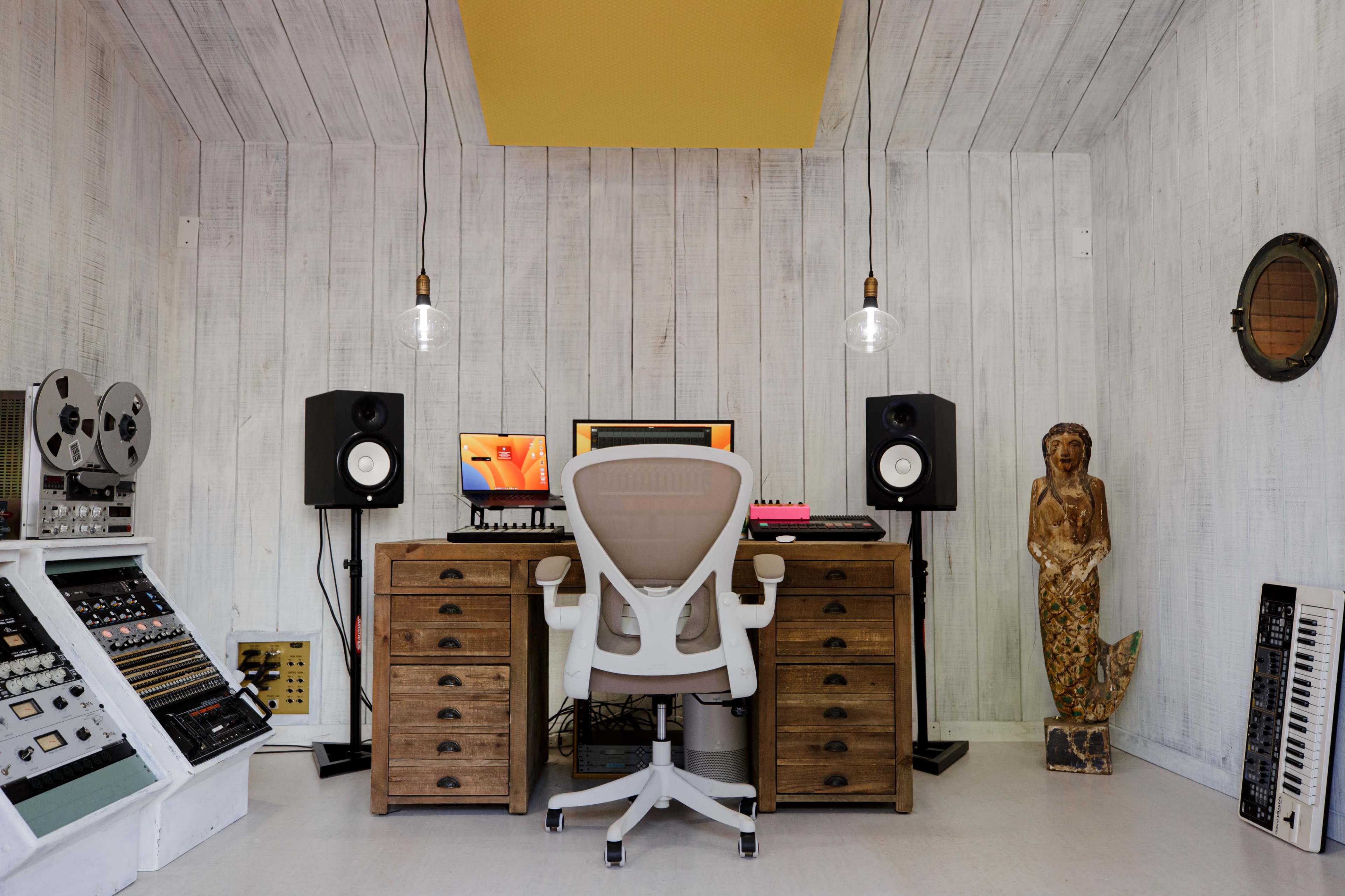 A music studio features a central wooden desk with a computer setup, flanked by speakers and vintage audio equipment, and a decorative statue in the corner.