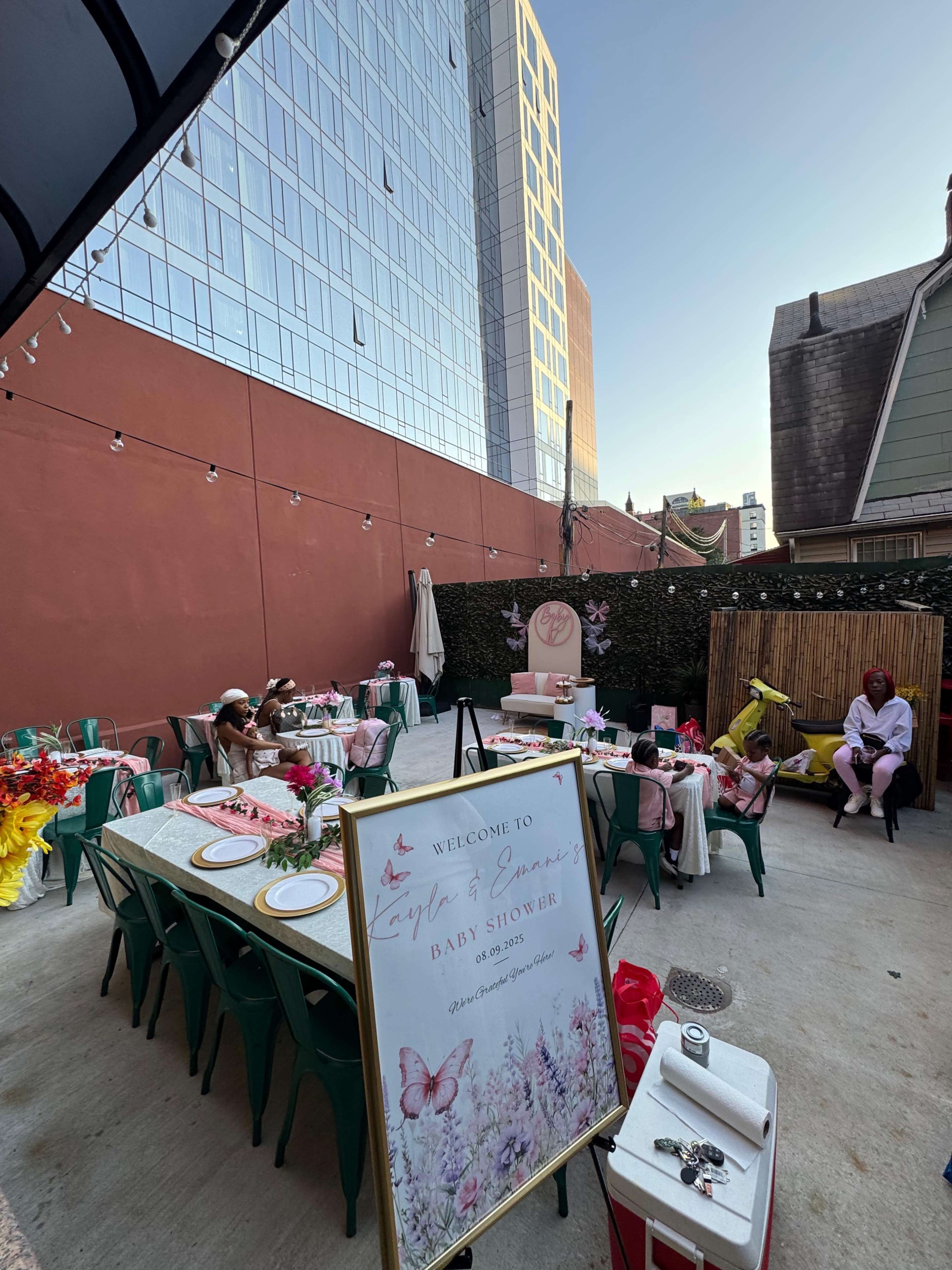 An outdoor baby shower setup is shown, featuring tables with decorations, a welcome sign, and guests seated in a courtyard surrounded by buildings.