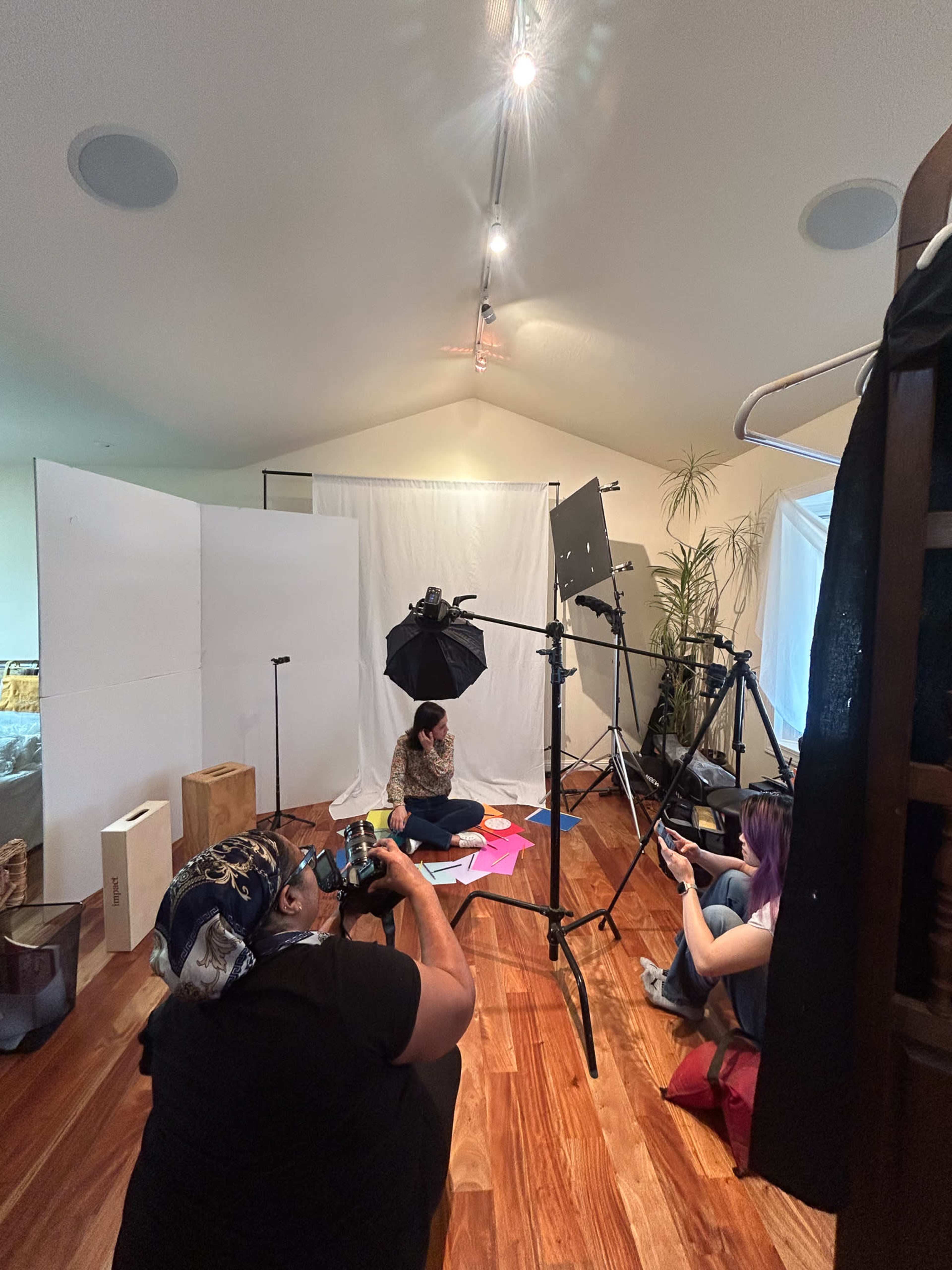 A photography setup features two individuals on the floor surrounded by colorful materials, with lighting equipment and backdrop panels arranged around them.