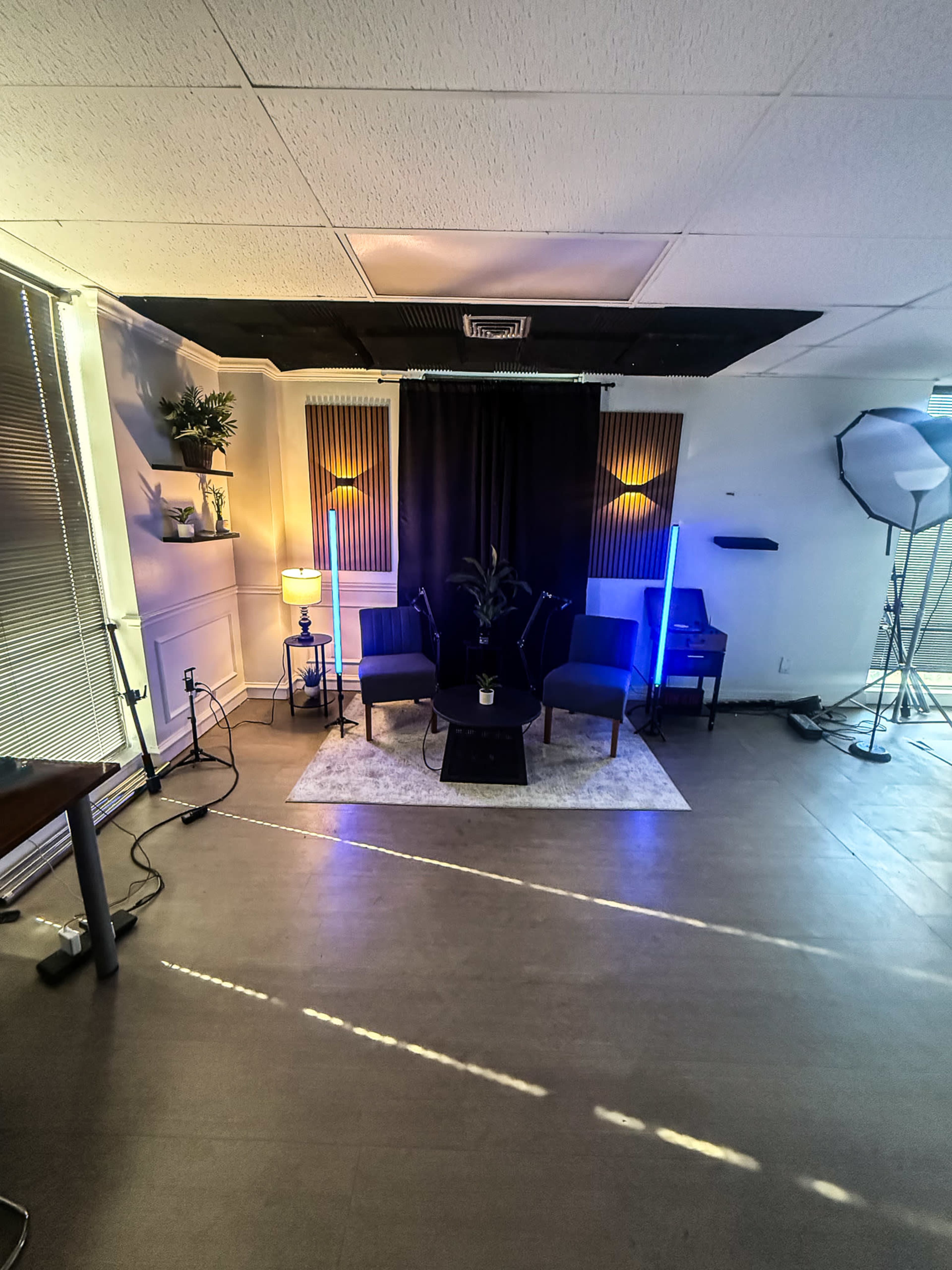 The image shows a modern interior space designed for a photo or video setup, featuring two blue chairs, a small table, a lamp, and a backdrop with lighting equipment.