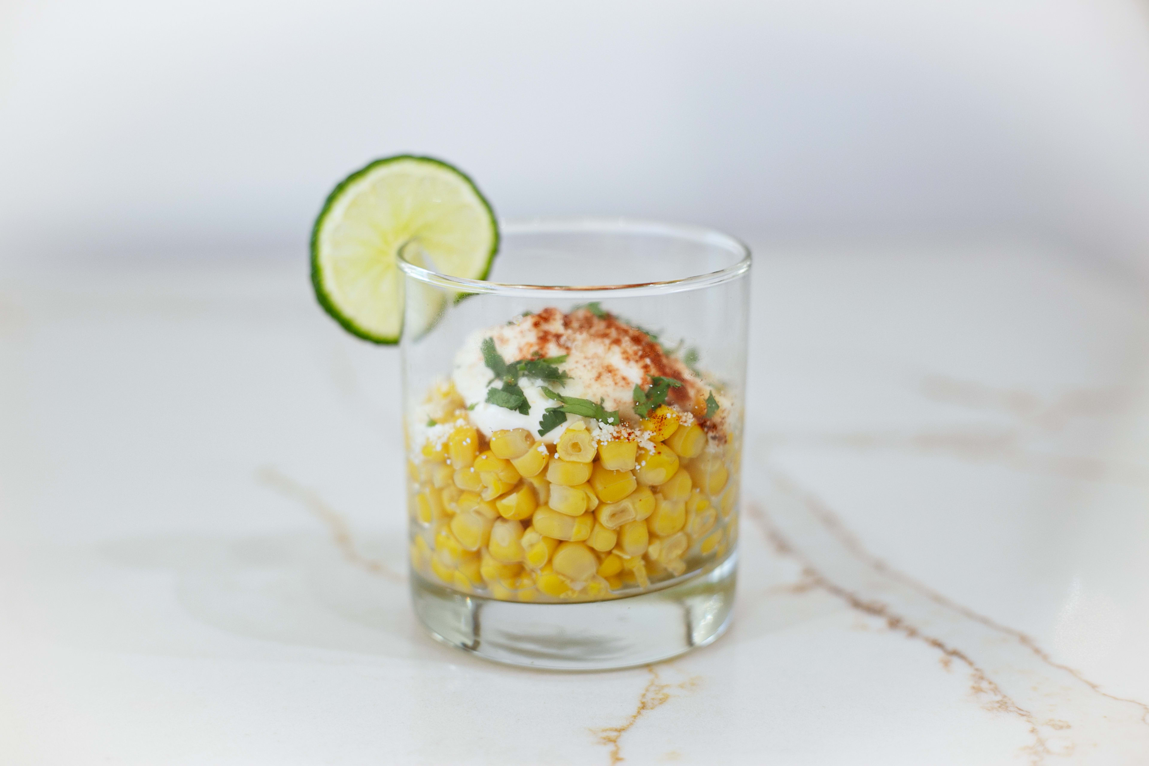 A glass filled with yellow corn topped with sour cream, paprika, and cilantro, garnished with a slice of lime.