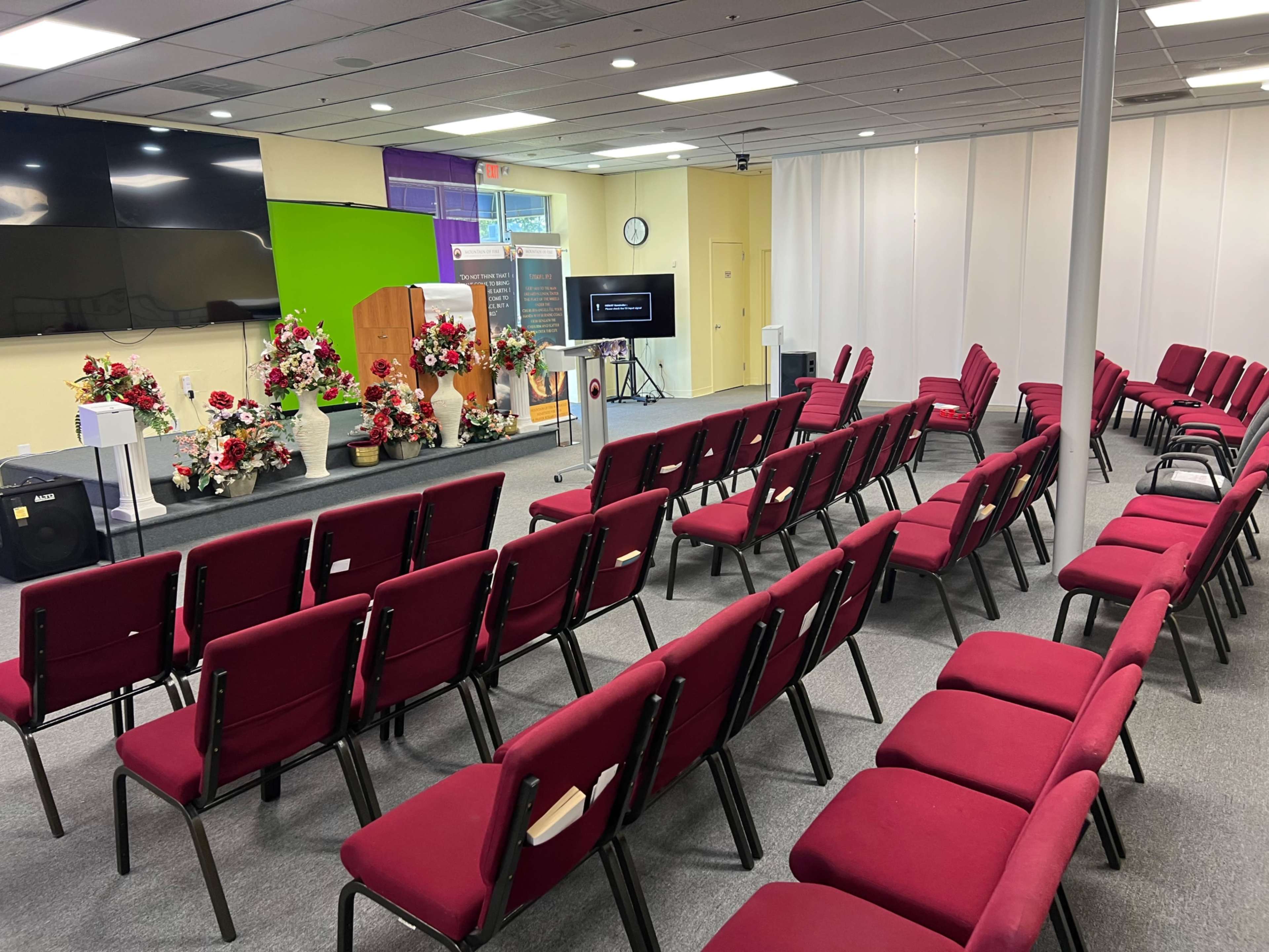 The image shows a set of rows of red upholstered chairs arranged in a spacious room with a podium, floral decorations, and a large screen at the front.