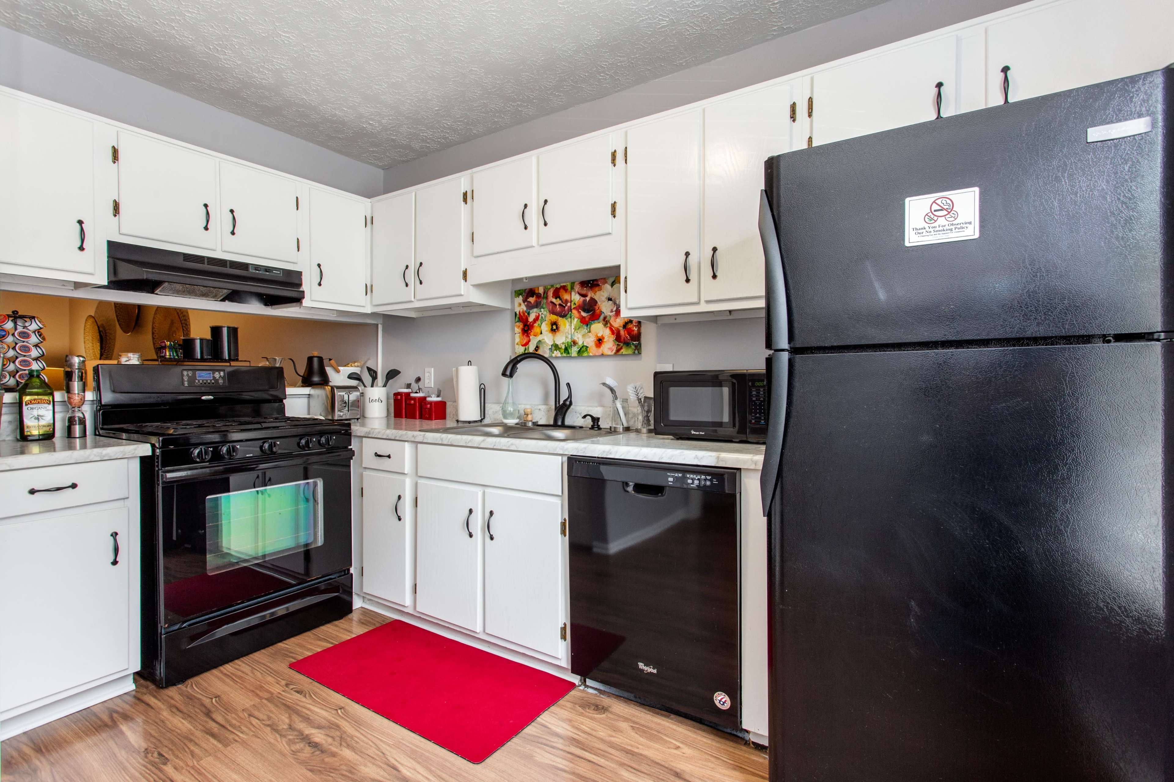 The kitchen features white cabinetry, black appliances, a red rug, and a floral wall decoration.