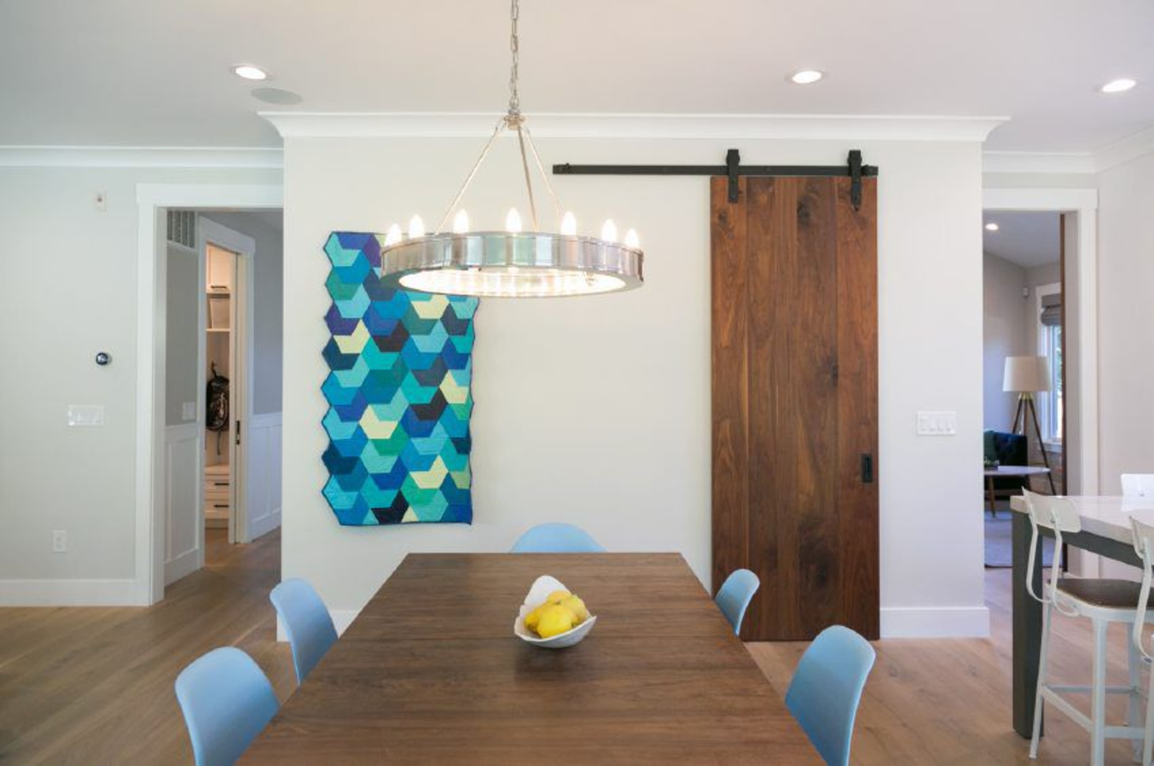 A dining area features a wooden table surrounded by blue chairs, with a large wall art piece in shades of blue and green and a sliding wooden door nearby.