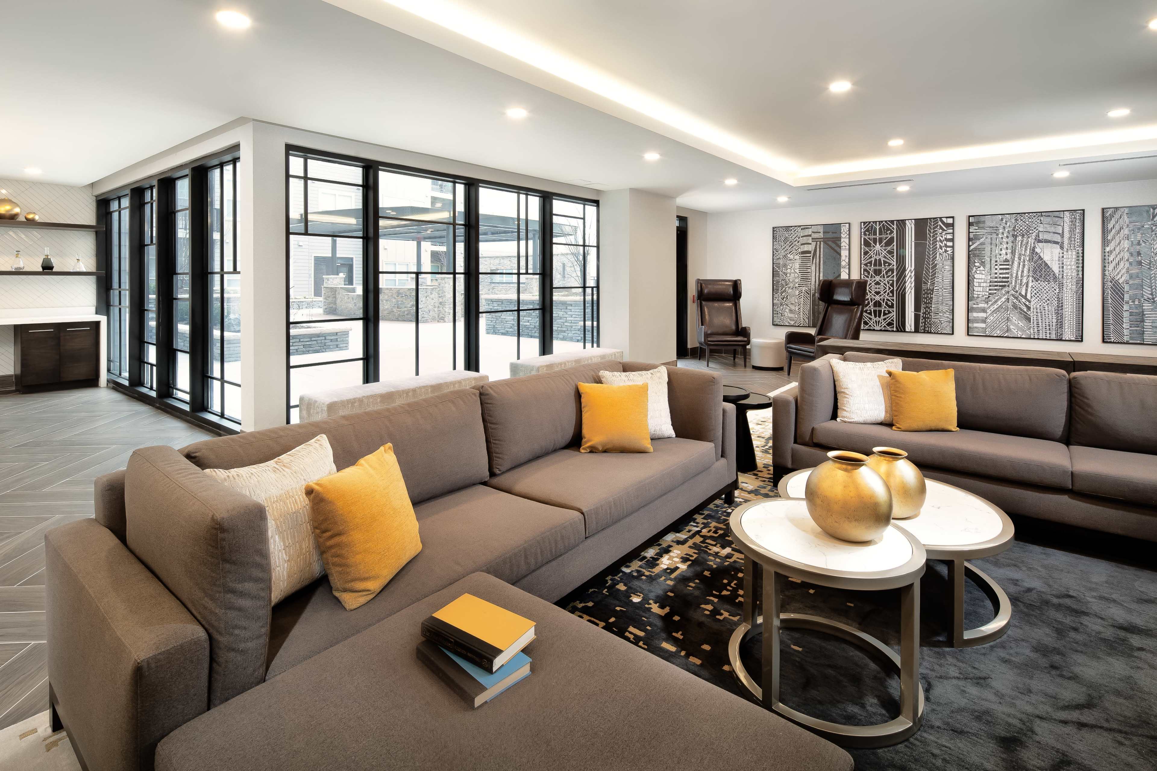 The image shows a modern living room featuring a large gray sectional sofa, two armchairs, a coffee table with decorative items, and large windows allowing natural light to enter the space.