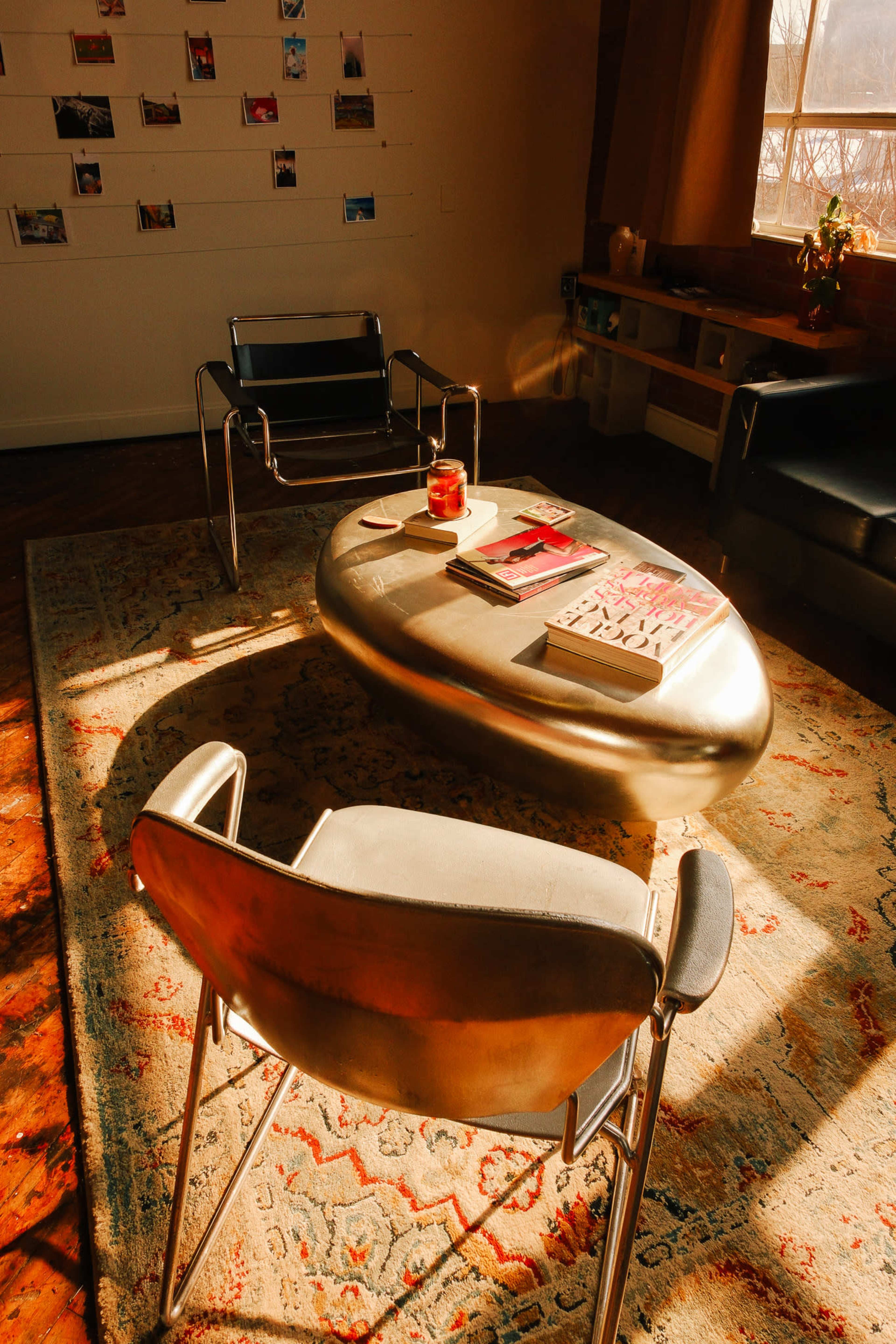 The image shows a cozy room with a shiny, oval coffee table surrounded by two modern metal chairs, illuminated by natural light streaming through a window.